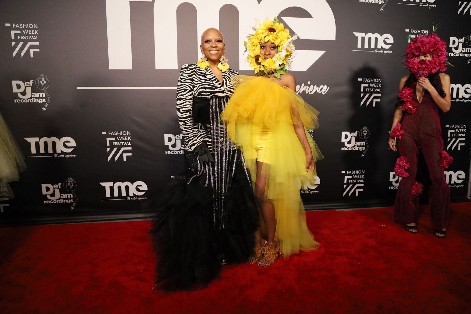Event guests pose in formal wear against black backdrop with TME logo at fashion show.