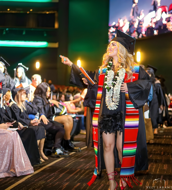 Graduate in decorative regalia with colorful serape stole celebrates during ceremony.