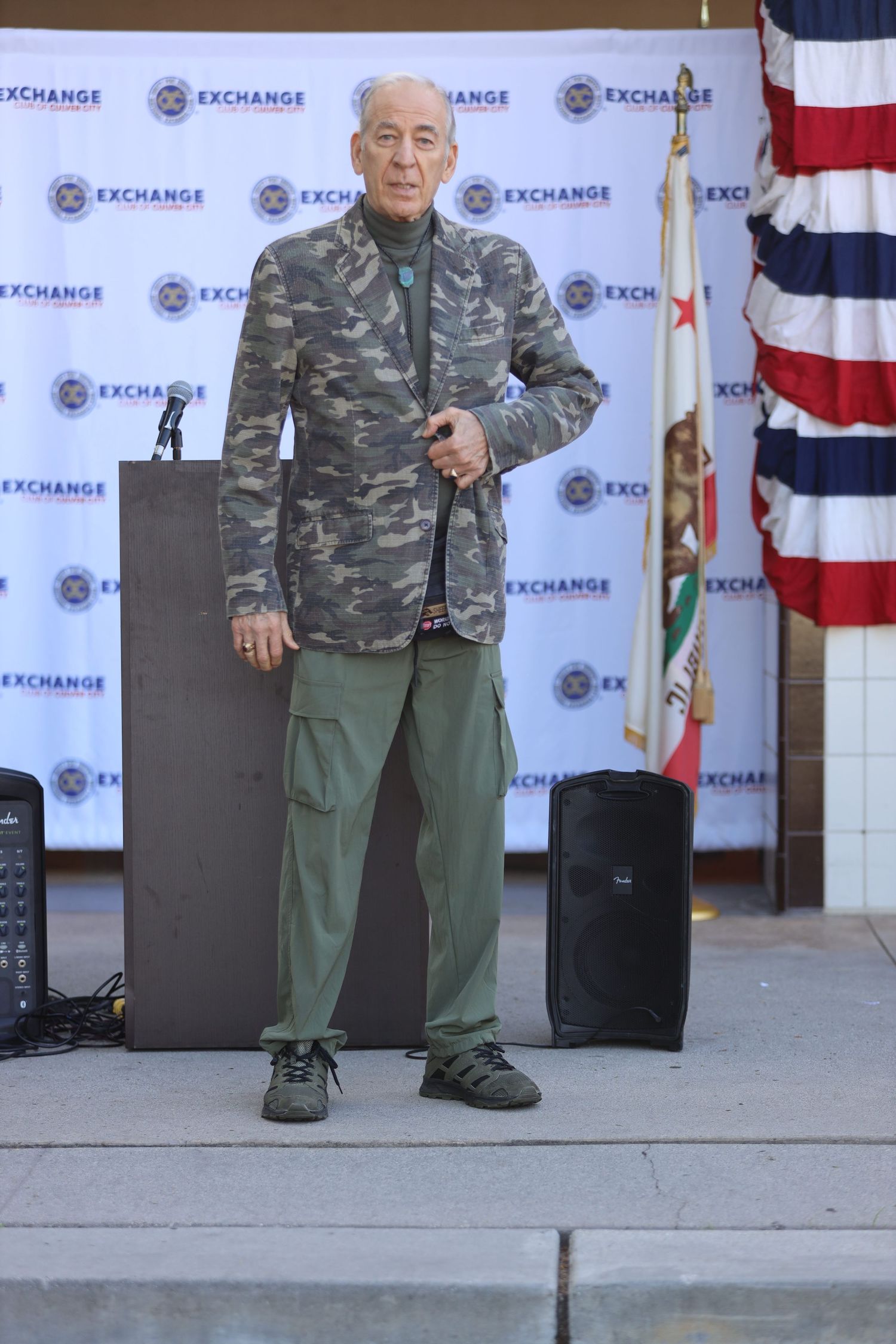 Senior man in camo jacket stands on stage at Exchange event with American flag display.