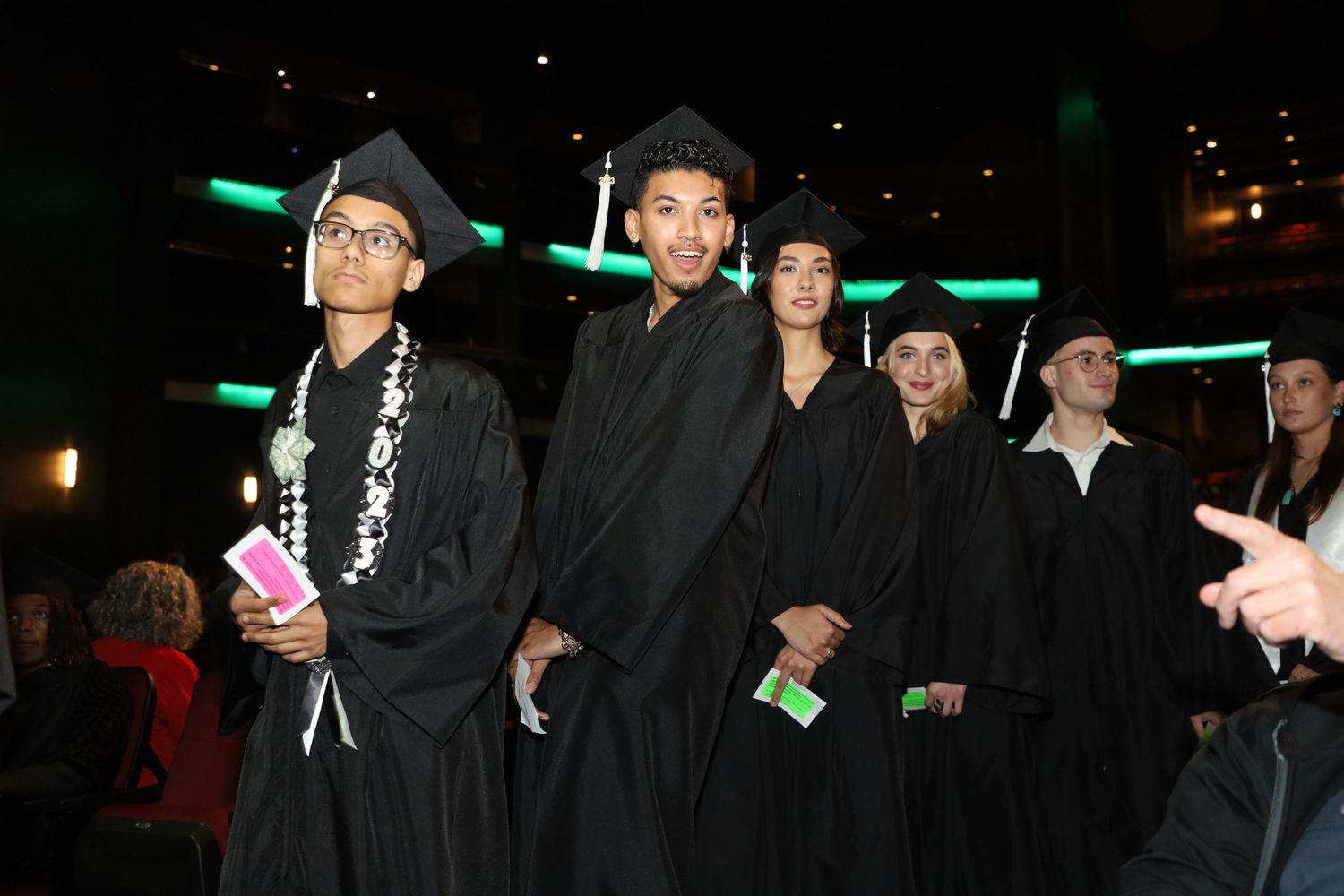 Row of graduates in black caps and gowns stand during ceremony under green lighting.