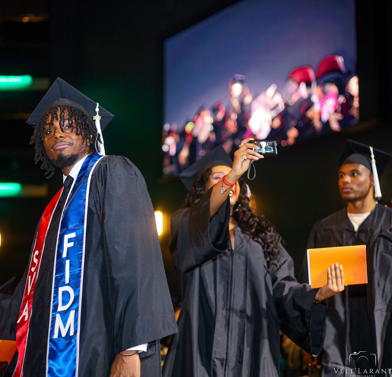 Graduate in cap and gown poses during graduation ceremony with projection screen in background.