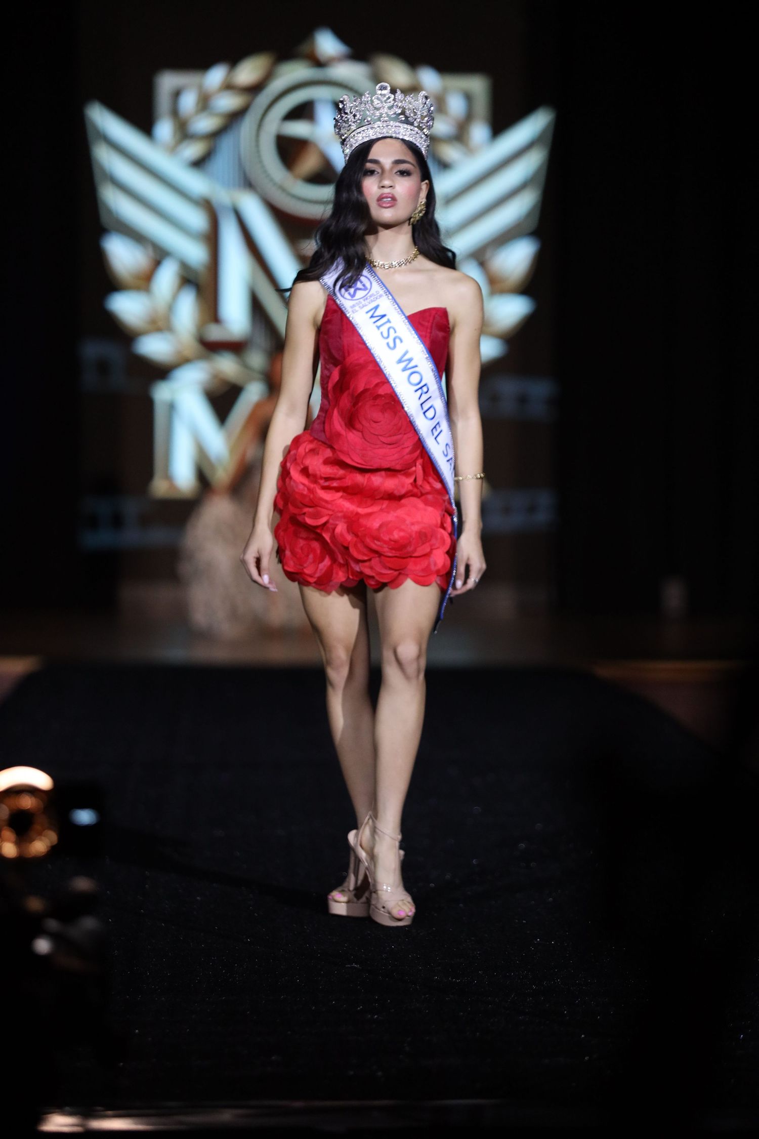 Beauty pageant winner poses in red cocktail dress and crown on dark runway with logo backdrop.