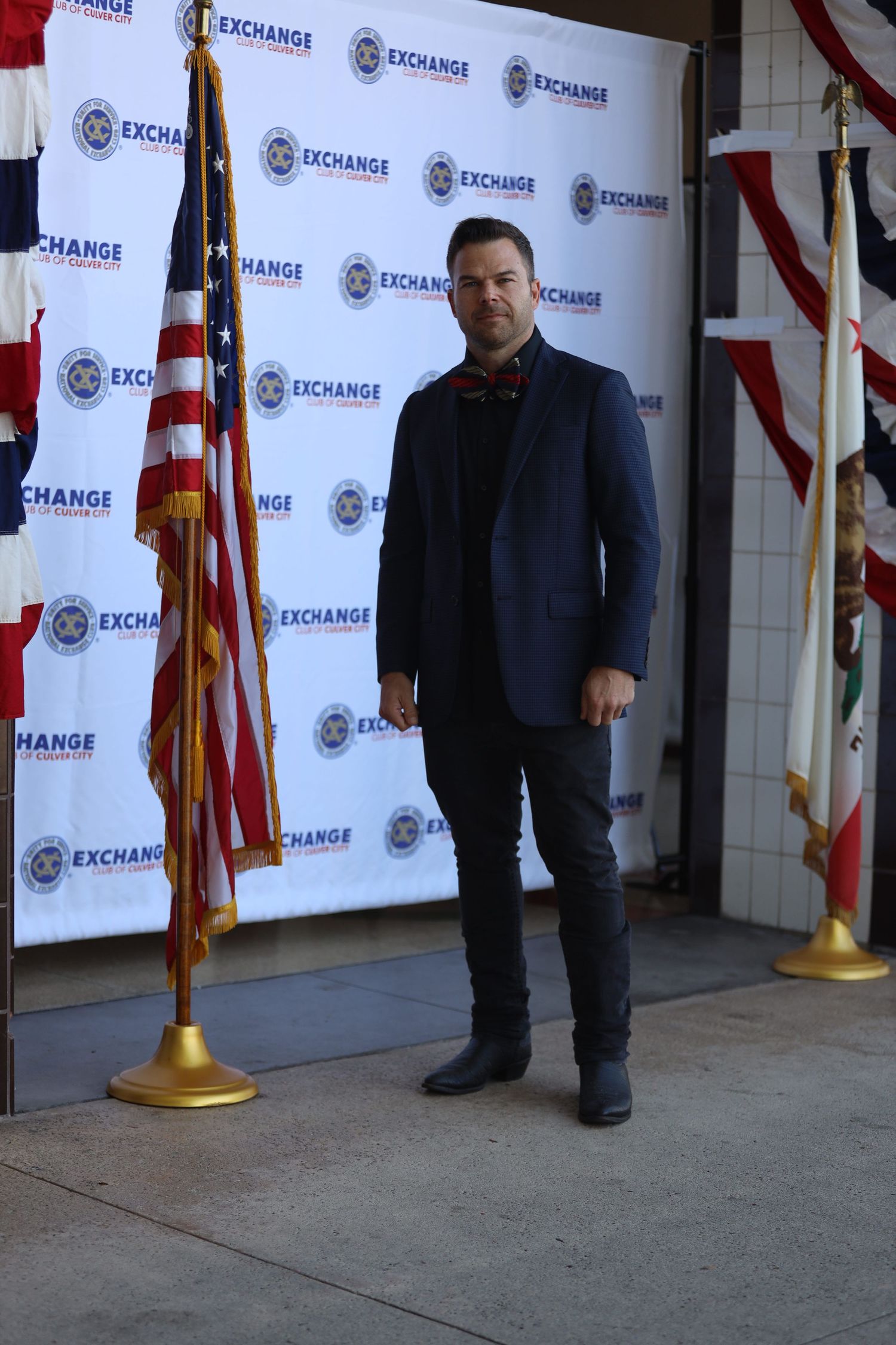 Figure in dark suit stands in front of step and repeat wall with Exchange branding.