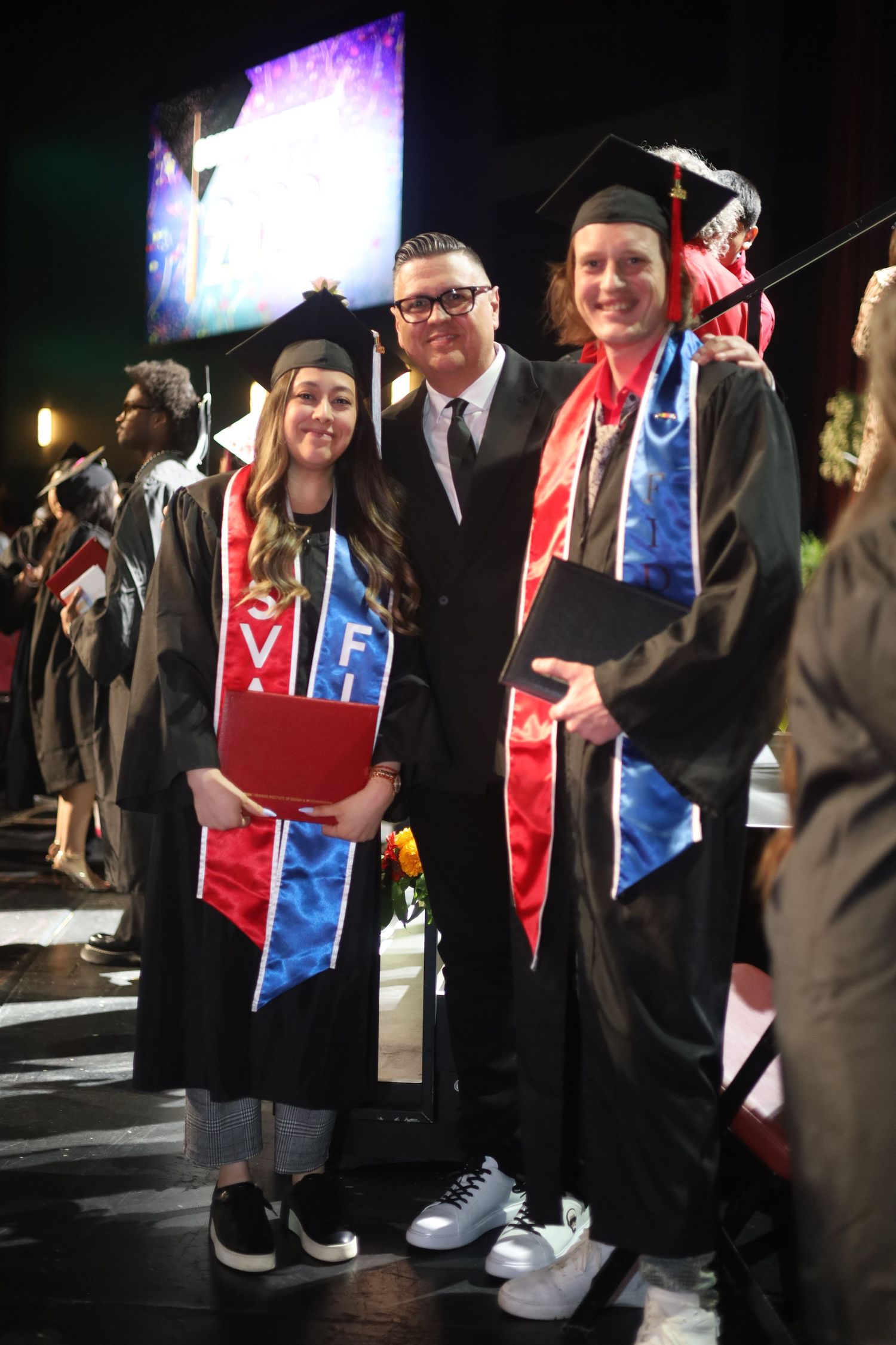 Graduates wearing red and blue stoles gather for photos at night.