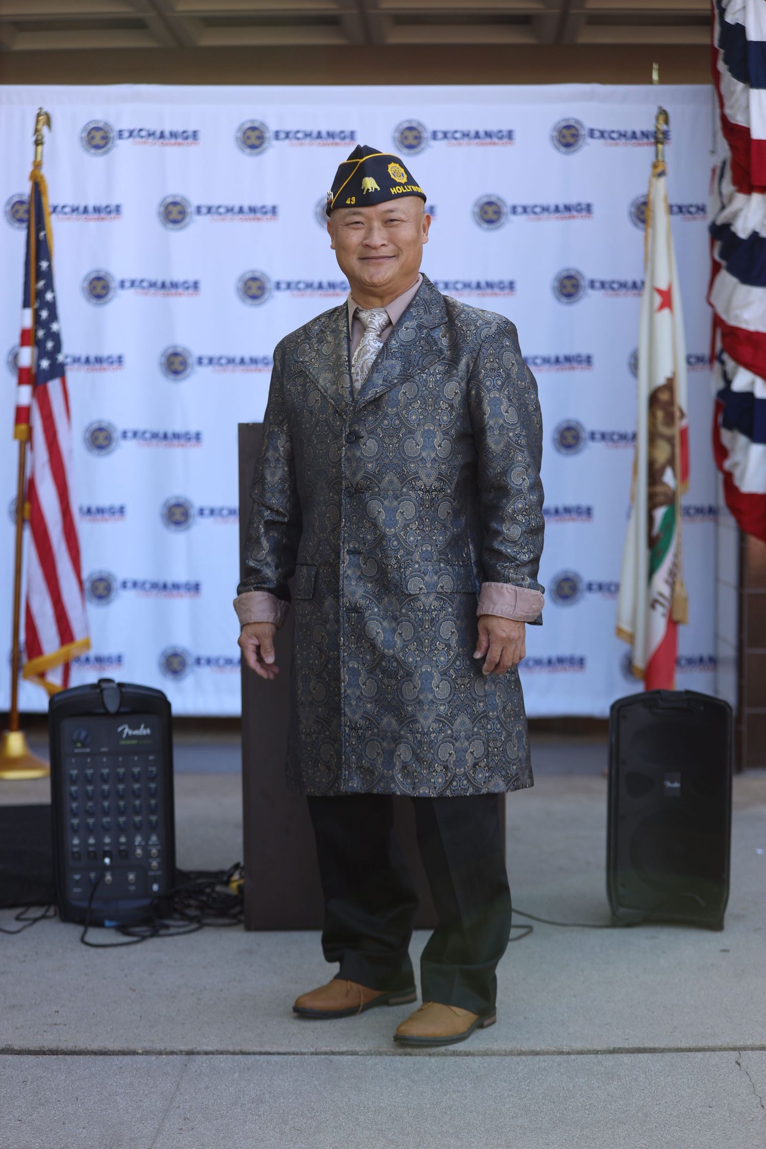 A person wearing a gray patterned jacket and dark pants stands in front of a branded backdrop with flags.