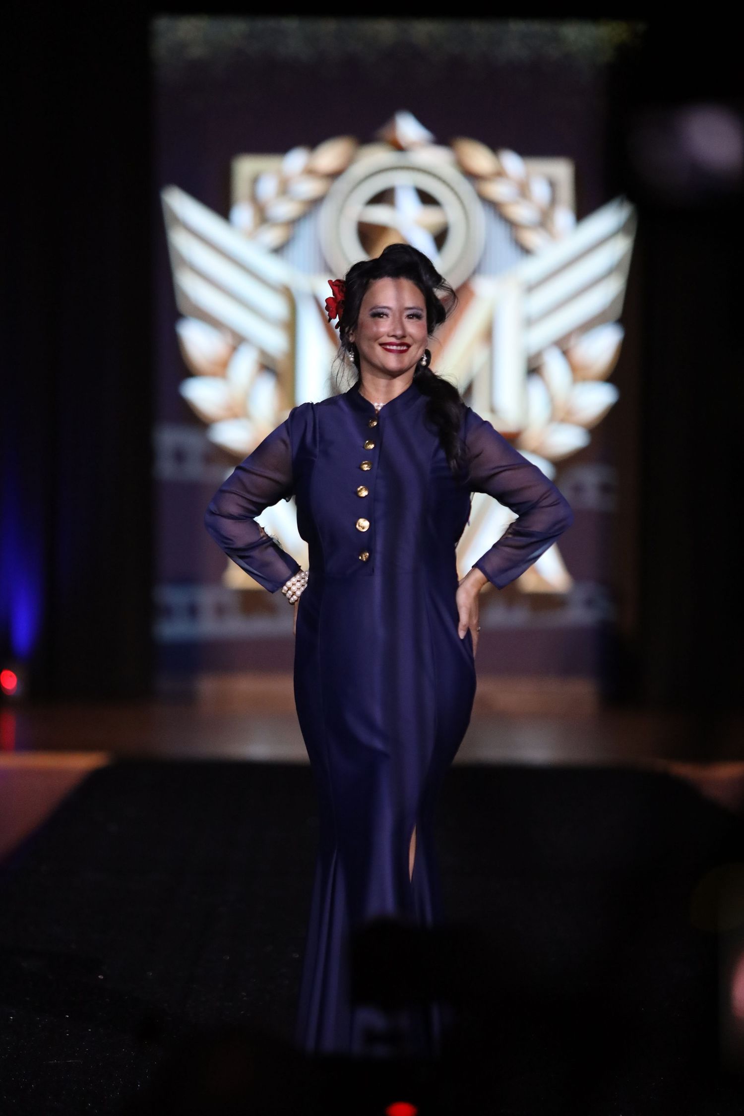 Model showcases navy military-style dress on dark runway with illuminated backdrop.