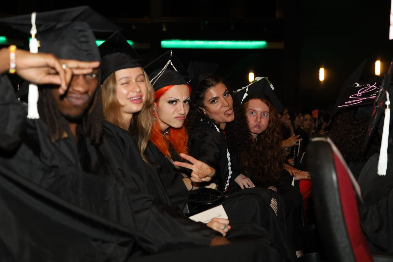Graduates in black caps and gowns gather during a dimly lit indoor graduation ceremony.