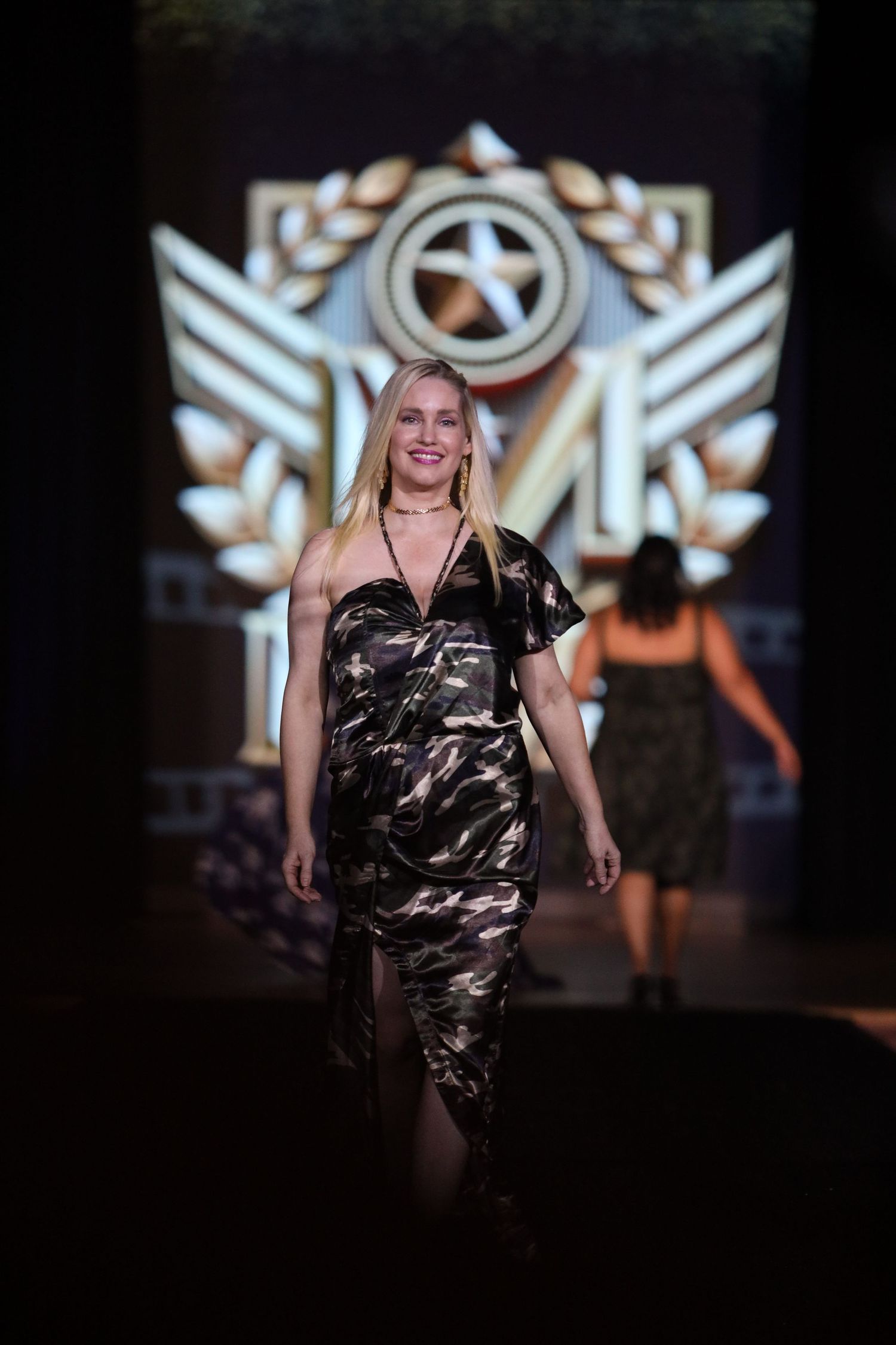 Model walks runway in camouflage print outfit with dramatic lighting and stage effects.