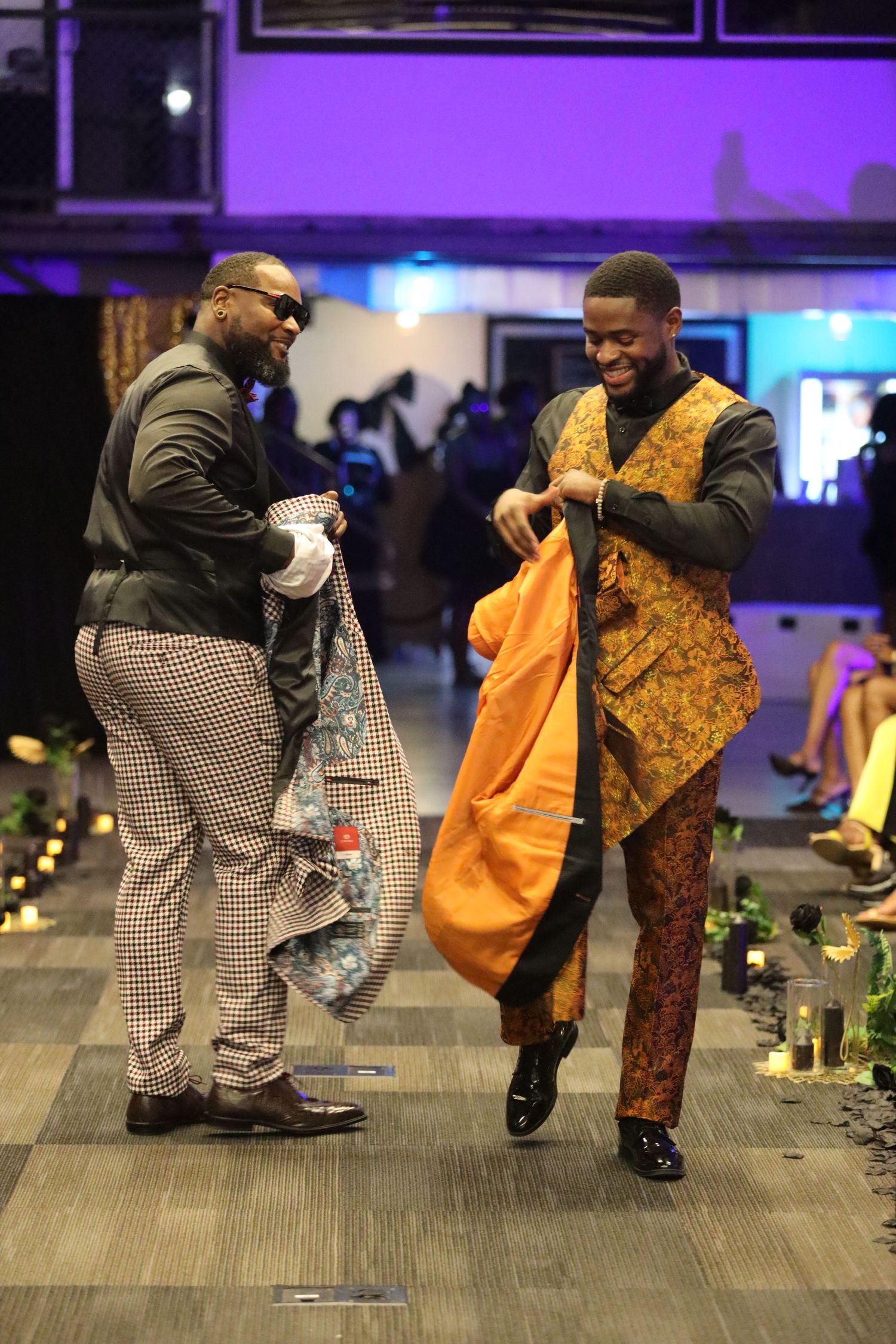 Two models interact while showcasing patterned suits on fashion runway.