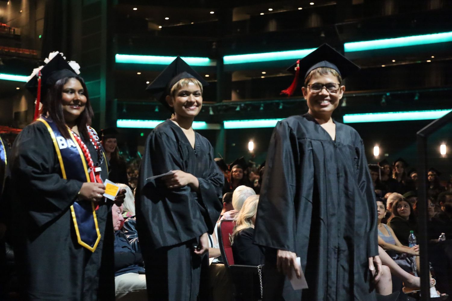 Graduates in black robes walking across stage during graduation ceremony.