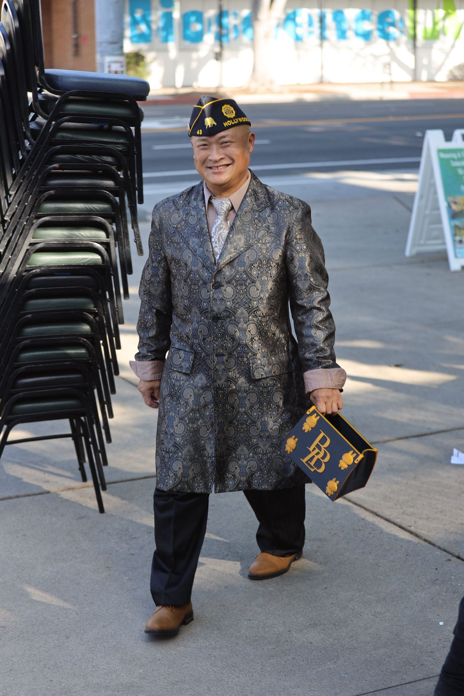 Veteran in patterned suit jacket and military cap walks past stacked chairs outdoors.