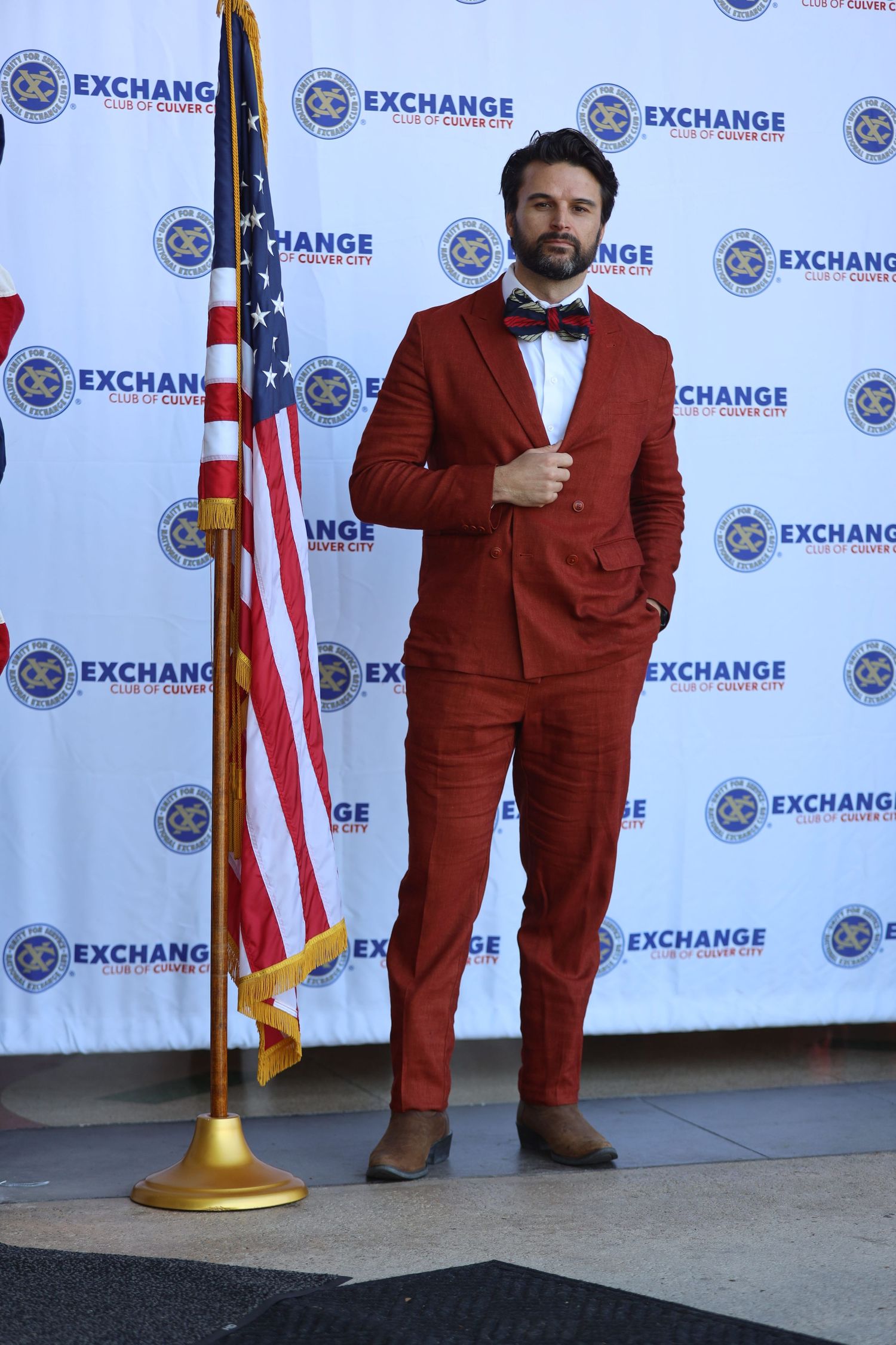 Person in a burgundy suit poses against a backdrop with Exchange logos and an American flag.