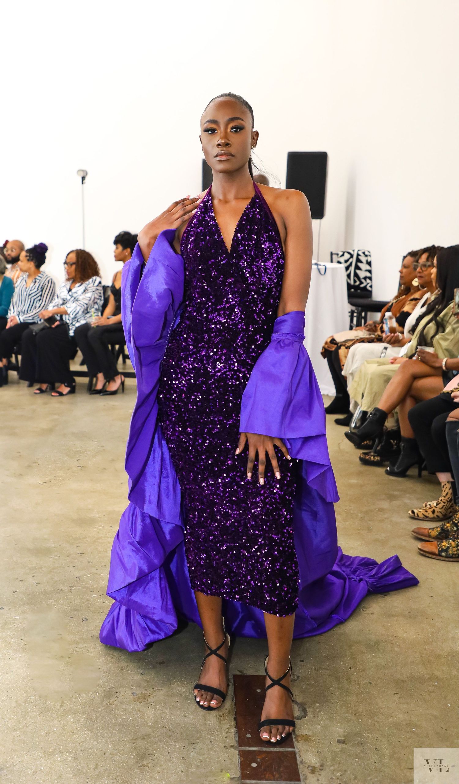 Model walks runway in sparkling purple sequin dress with dramatic flowing cape at fashion show.