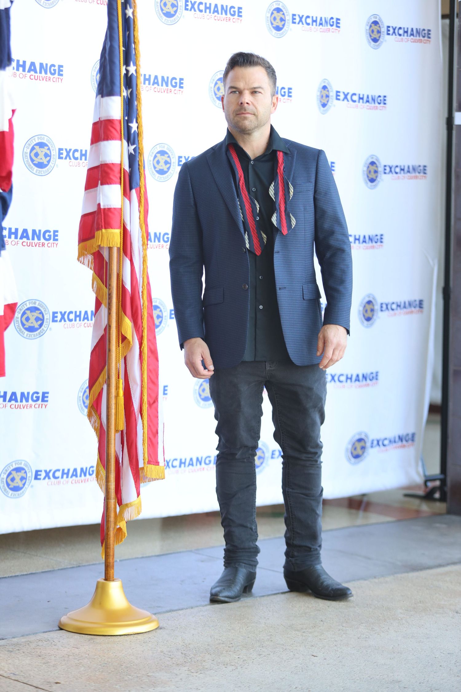 Person in dark jacket stands near gold flag stand against branded backdrop.