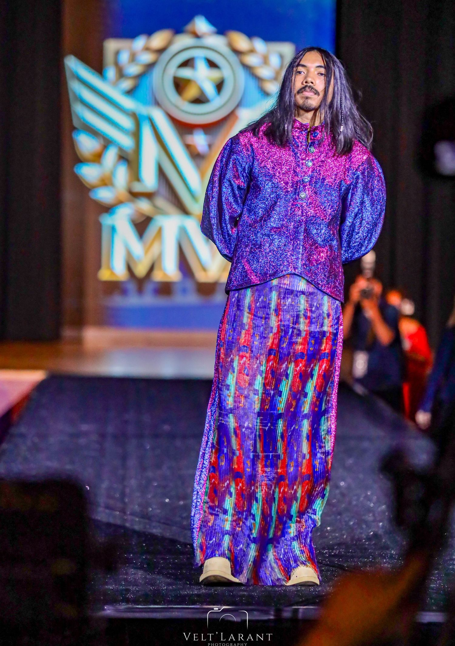 Fashion model in vibrant purple patterned outfit walks the runway against illuminated logo backdrop.