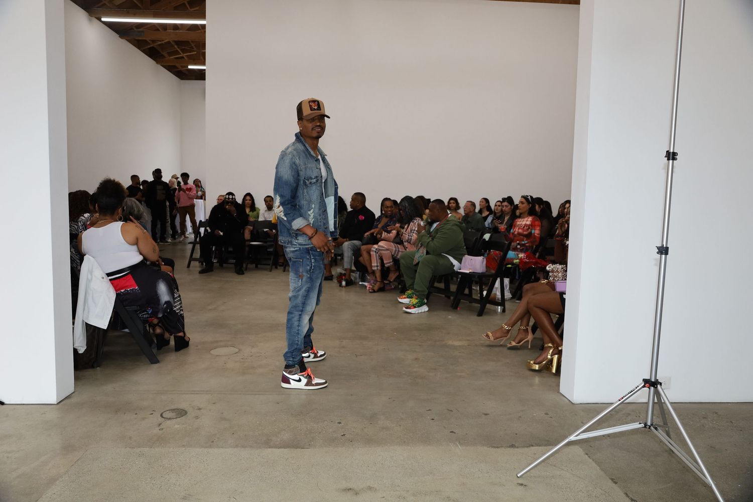 Model walking down a runway in casual denim outfit with audience seated along both sides.