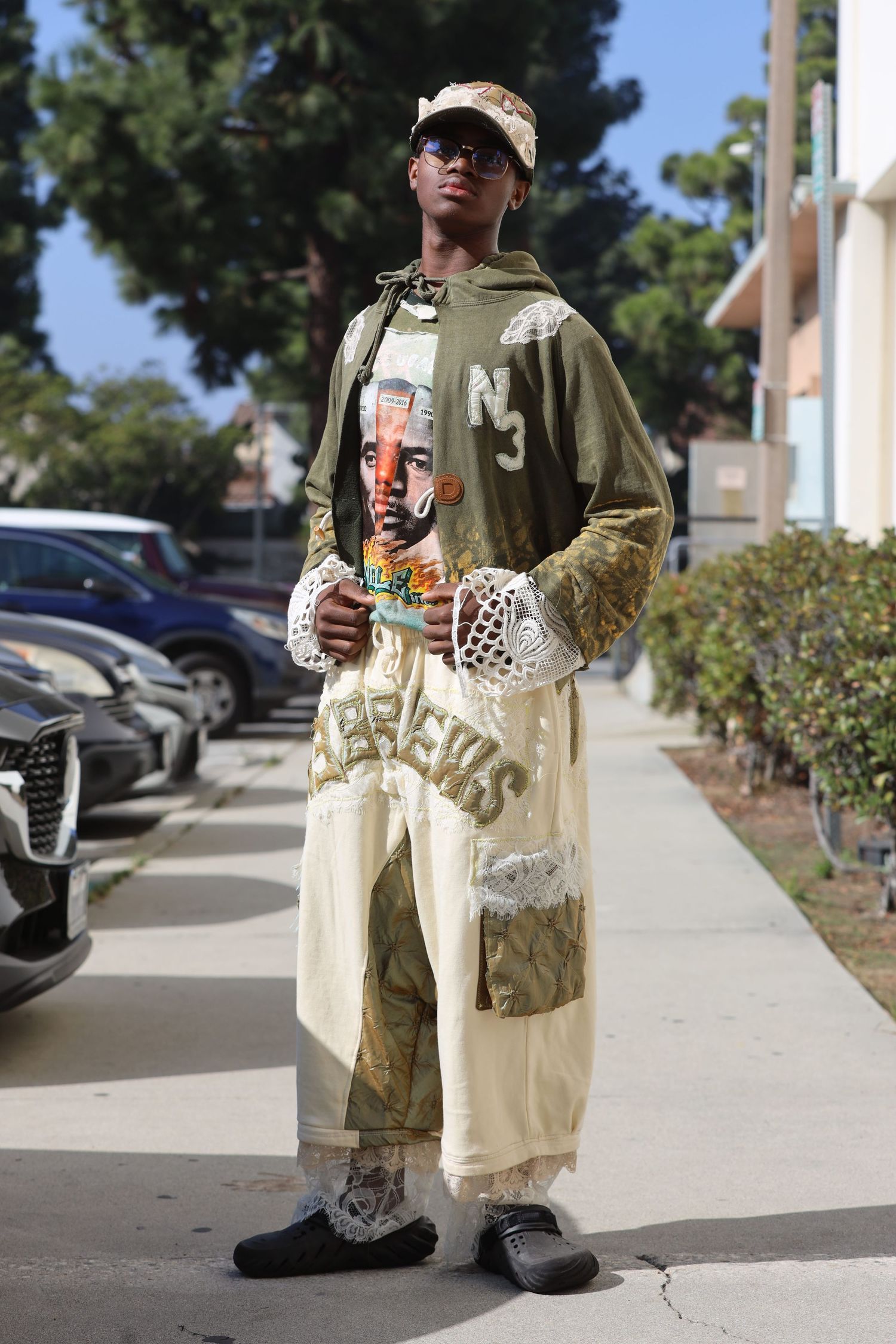 Person in streetwear poses outdoors wearing olive jacket, graphic tee, and cream-colored pants.