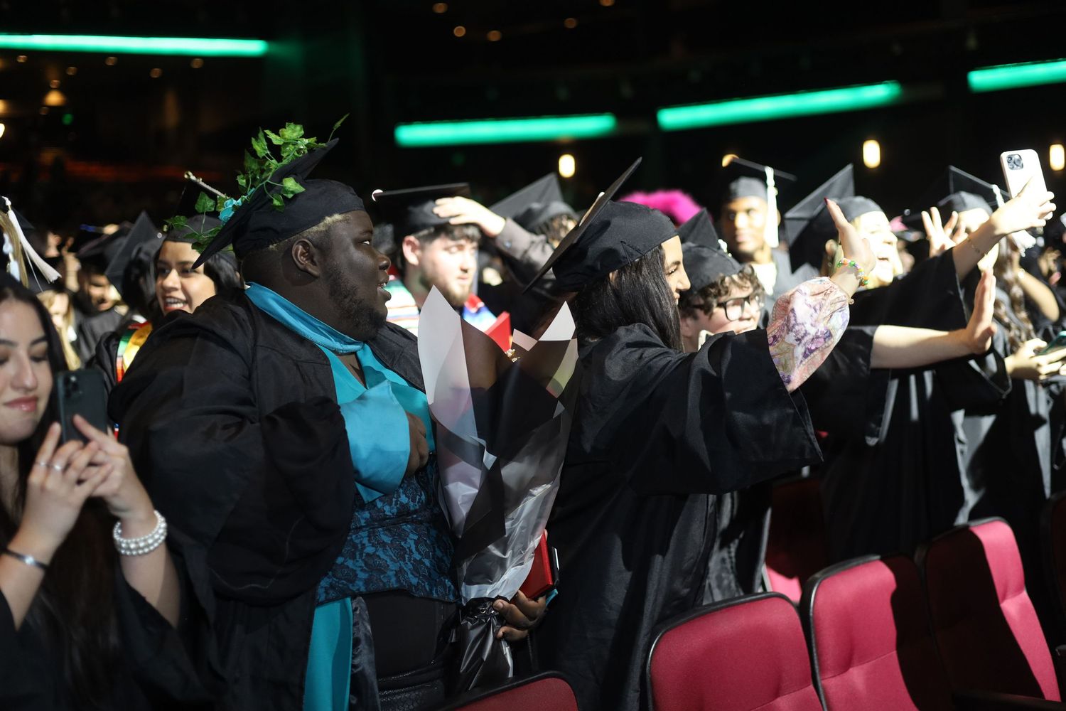 Graduates in black caps and gowns celebrating at a commencement ceremony with bright lighting overhead.