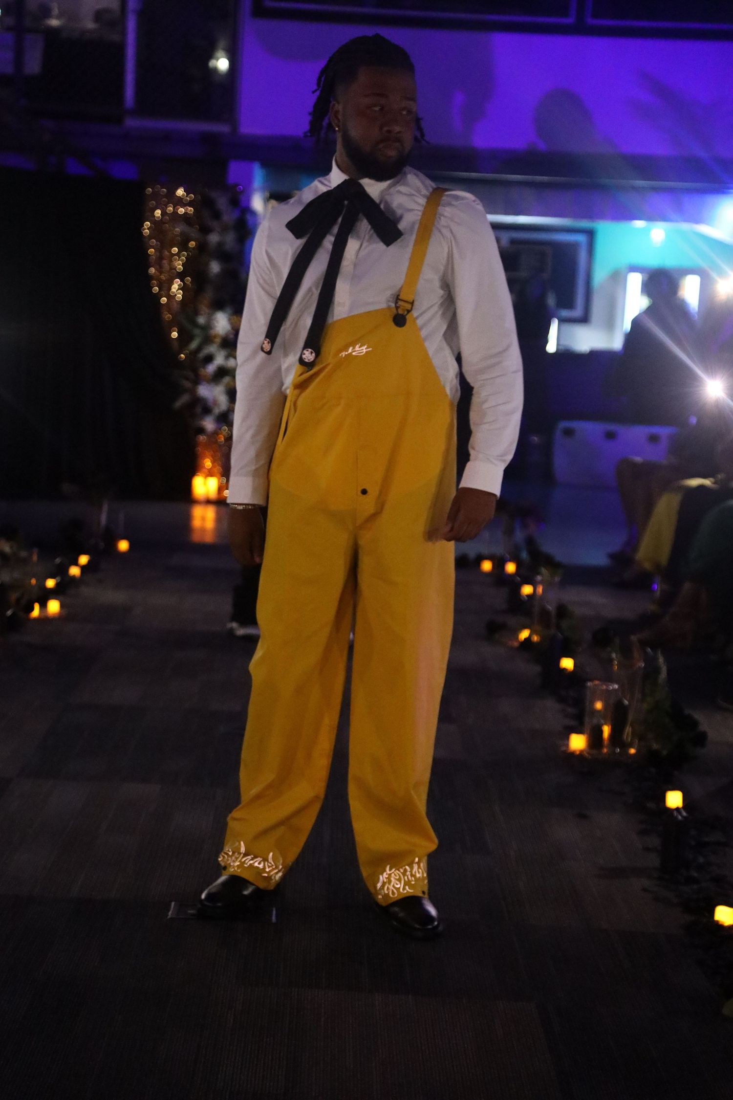 Person wearing bright yellow overalls and white shirt in dimly lit venue with purple lighting.