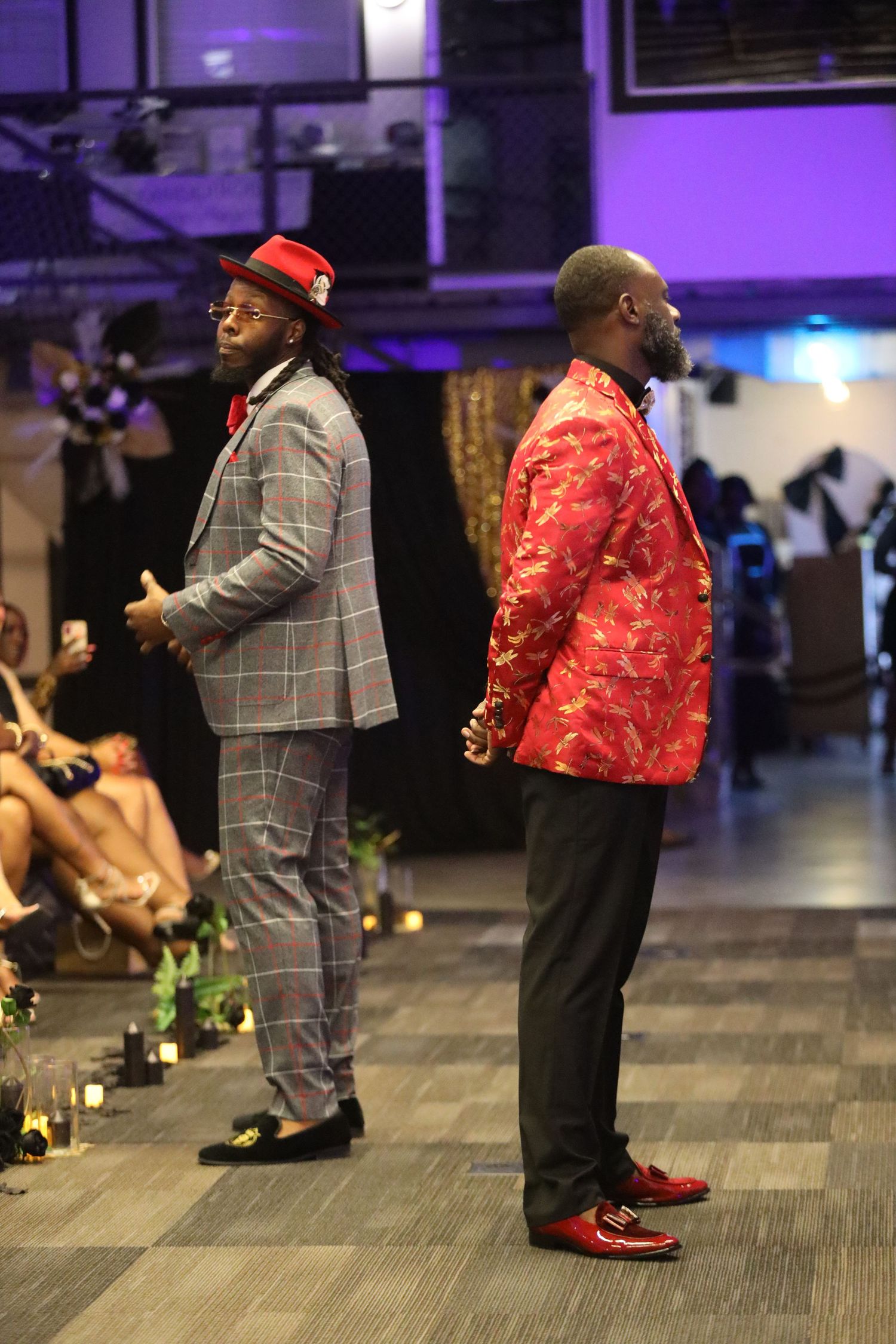 Models present plaid and red patterned suits on fashion runway with purple ambient lighting.