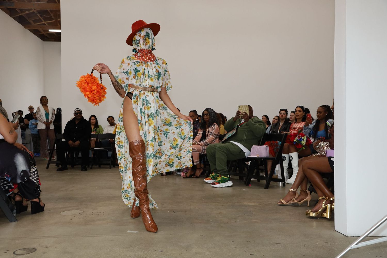 Model struts down catwalk wearing vintage-inspired floral dress and red accessories.