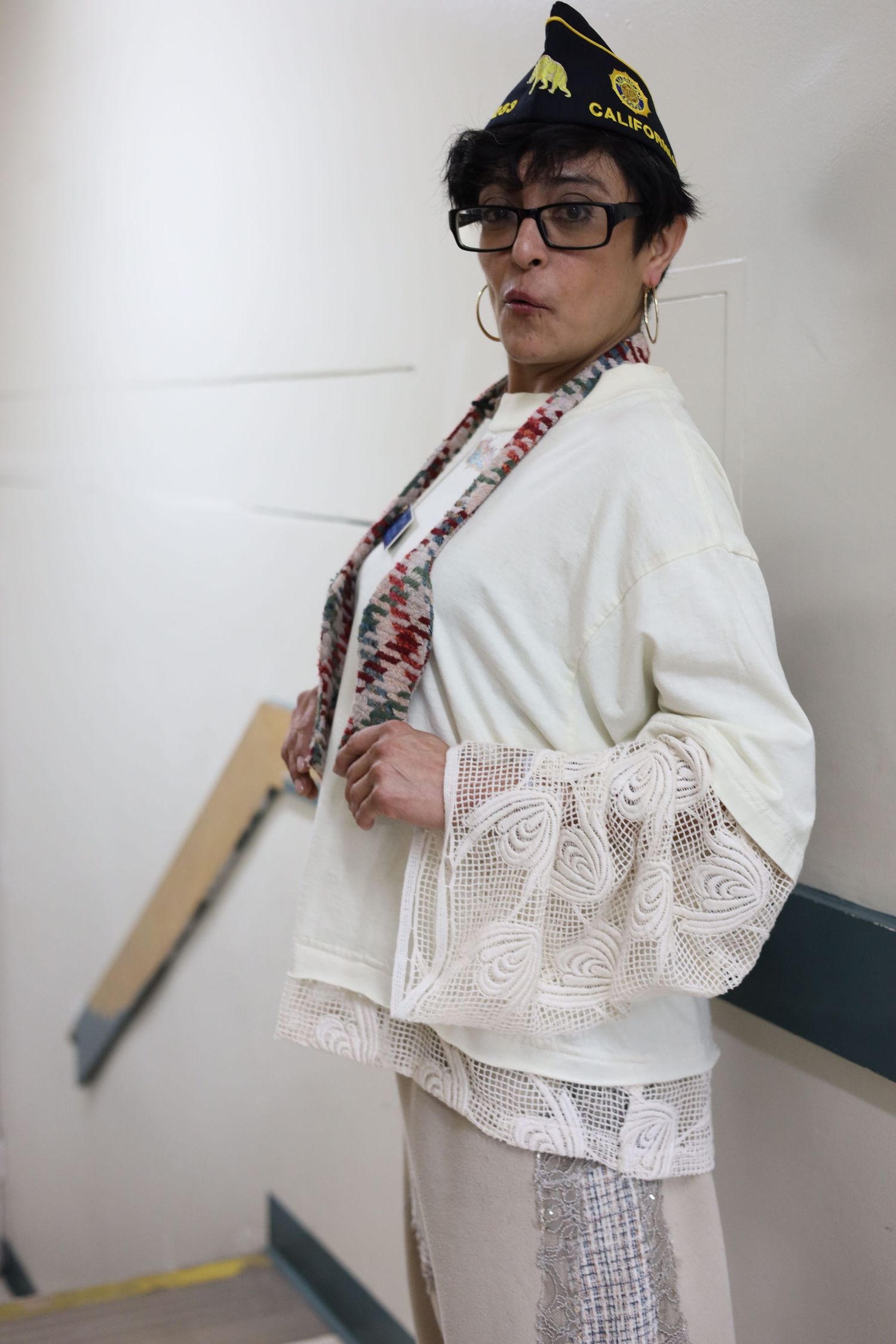 Person wearing a white lace top and dark hat poses against a white wall.