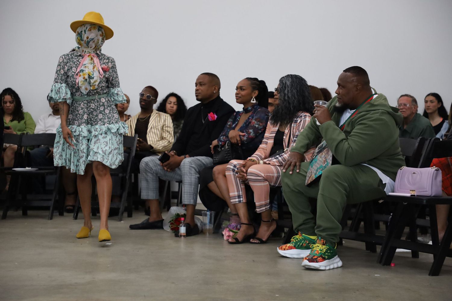 Fashion show models present designs on a concrete runway with audience seated along sides.