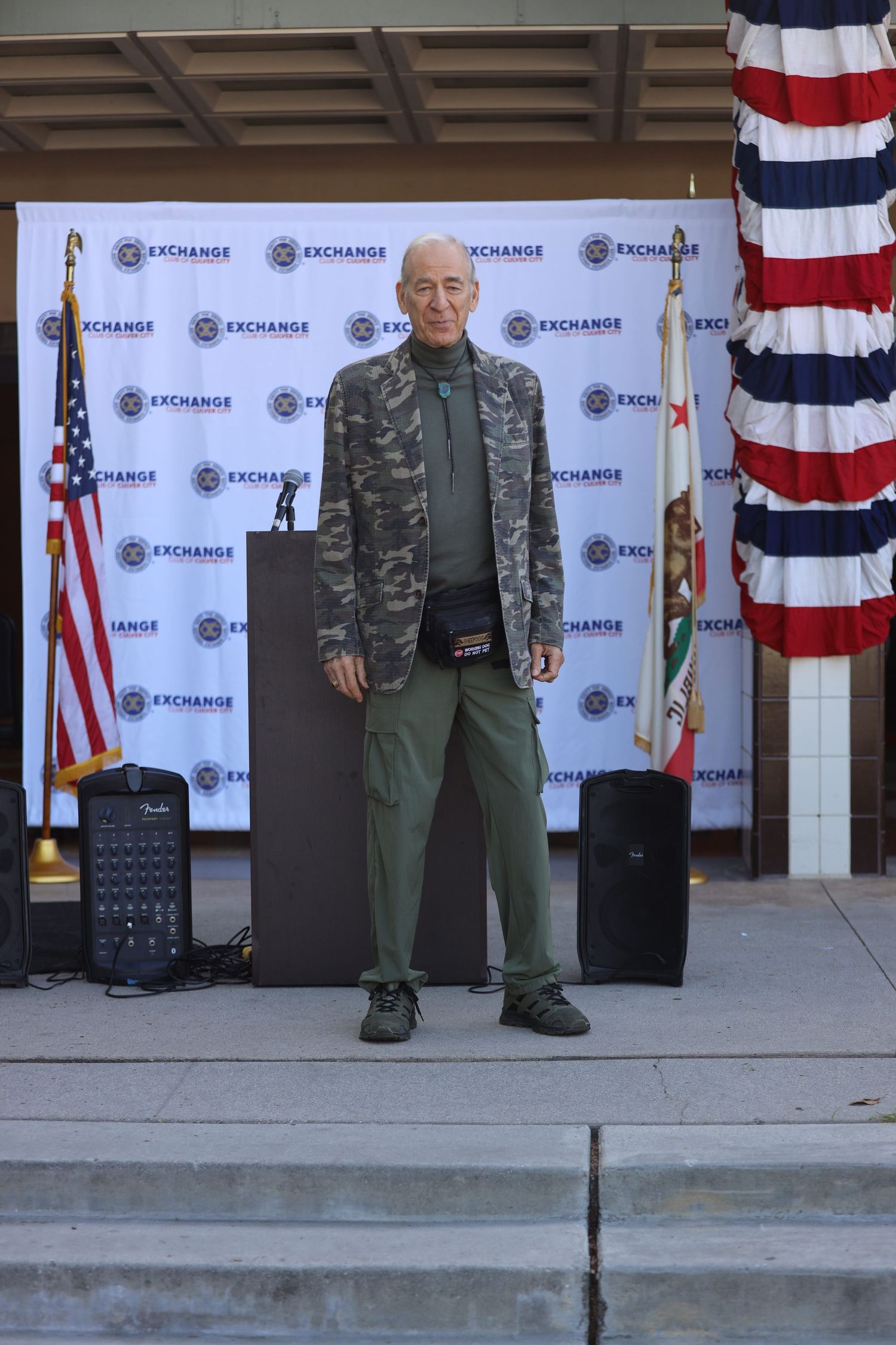 Senior man in camouflage jacket poses at military event with American flag backdrop.