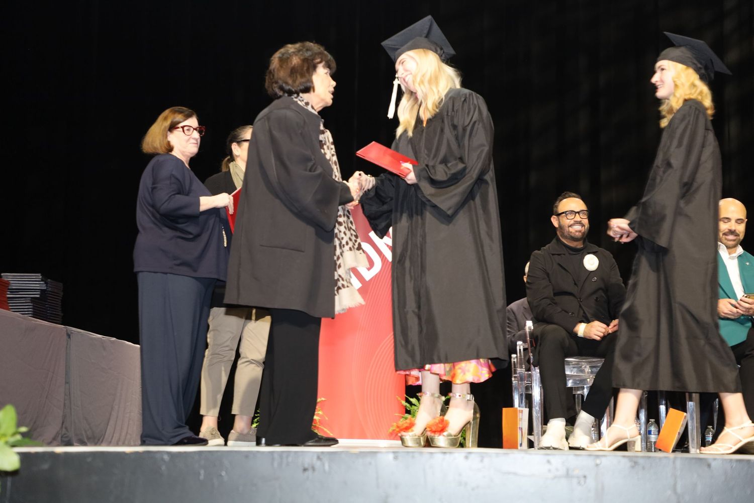 Students in black graduation gowns receive diplomas on stage at commencement.