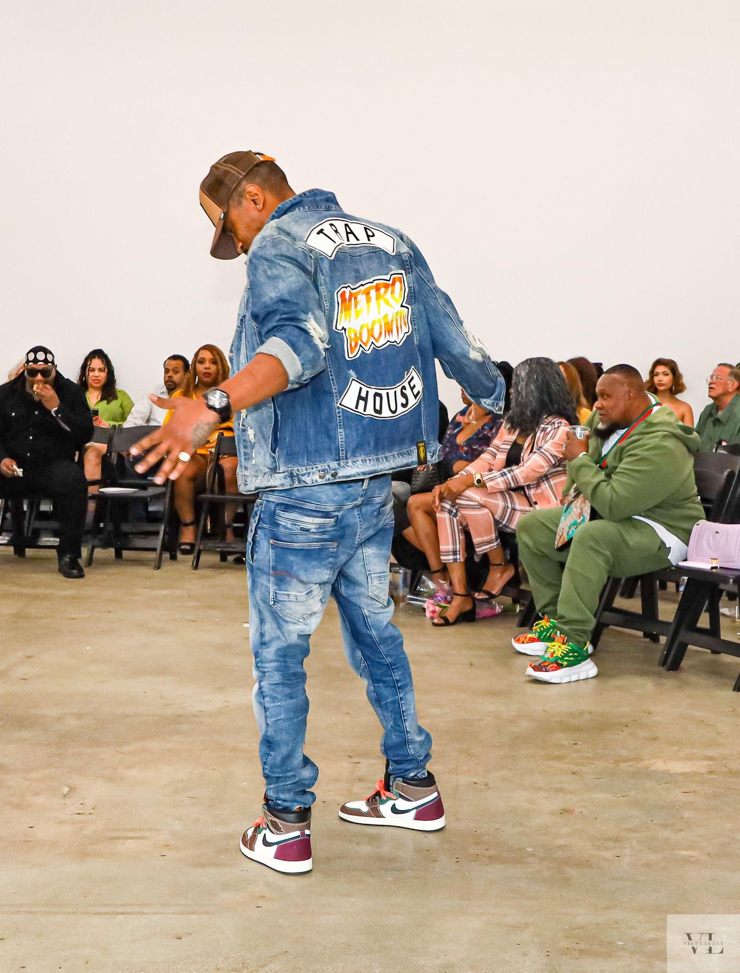 Performer in denim jacket and jeans dancing at an indoor fashion show with audience seated behind.