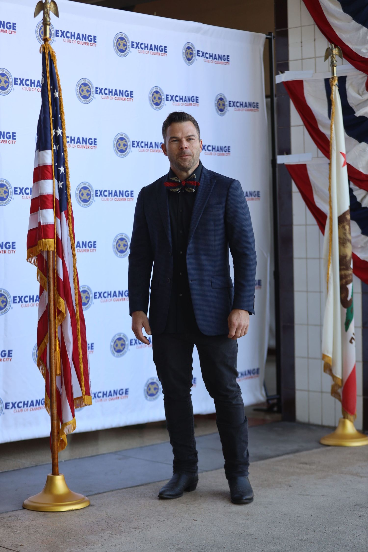 Figure poses in dark suit against branded backdrop with American flags.