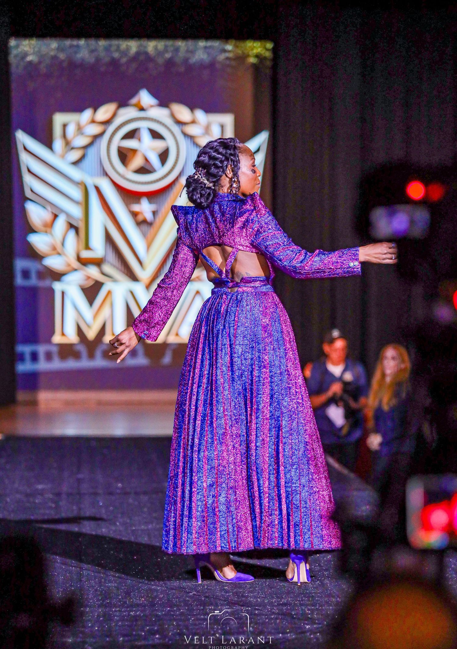 Model wearing a sparkly purple gown performs on stage with dramatic lighting and backdrop.