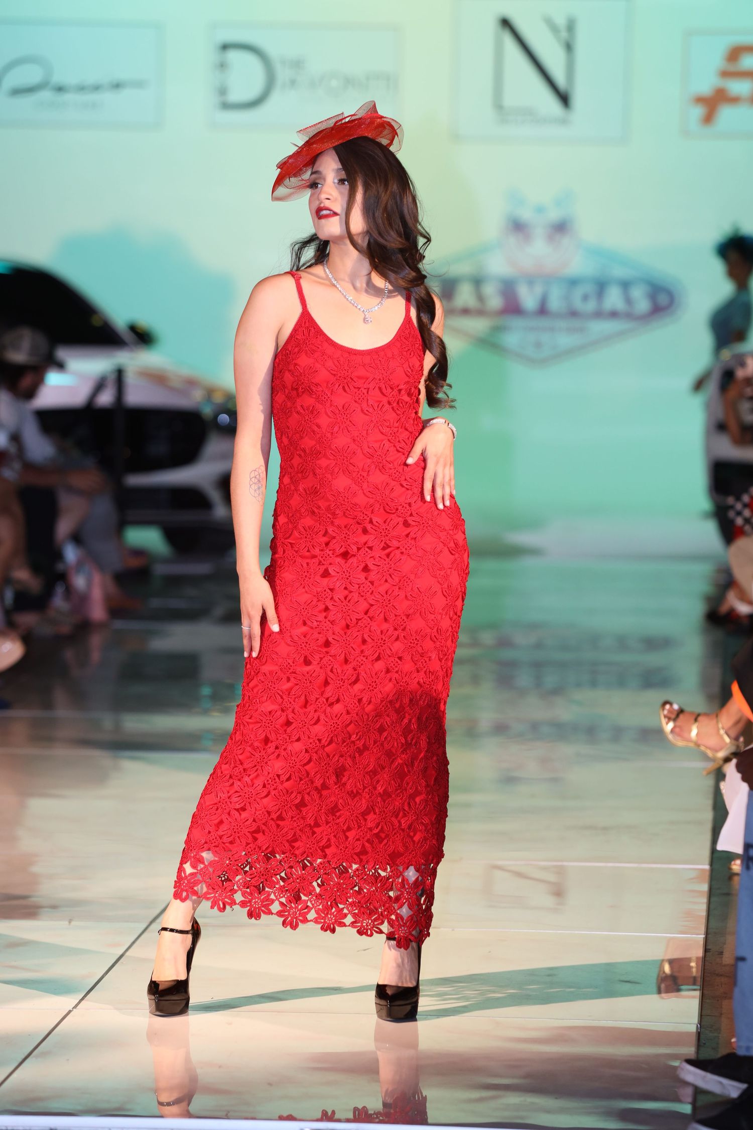 Model showcases red lace maxi dress with matching headpiece on fashion runway.
