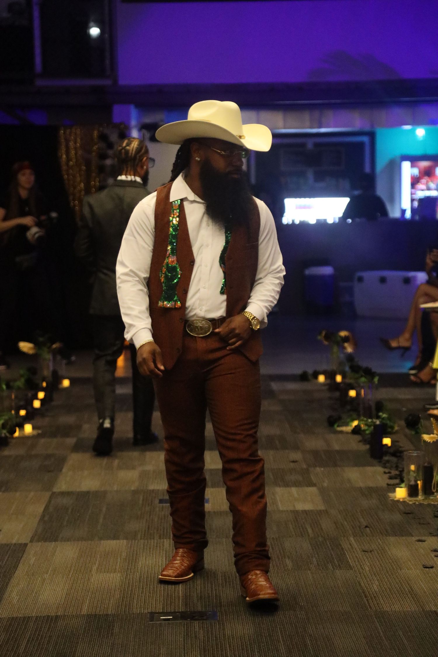 Person in western attire with cowboy hat and brown vest walking across carpeted floor.