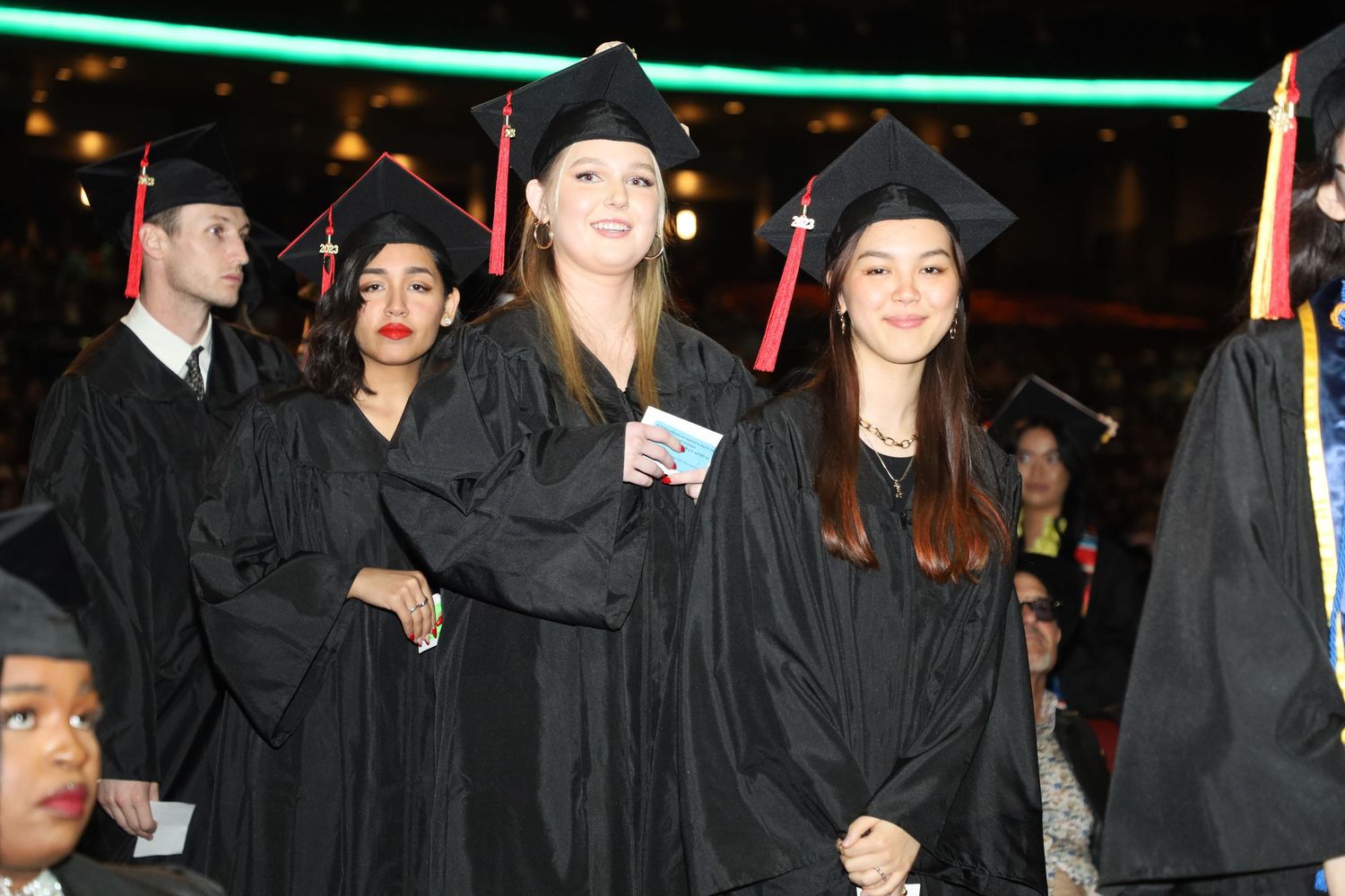 Row of graduates in black academic regalia standing during indoor ceremony.