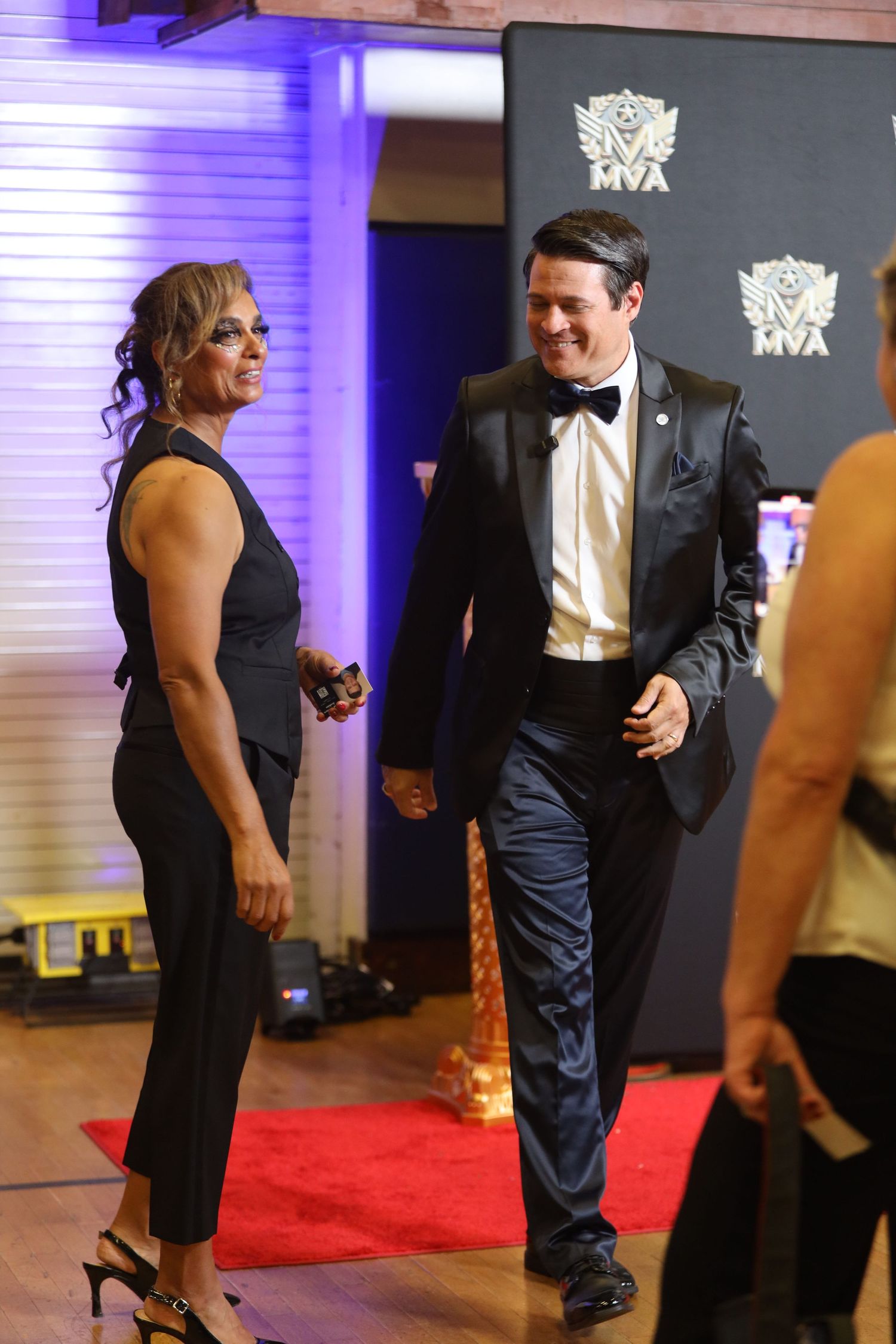 Formal event attendees in black tie attire interact on red carpet with purple lighting.