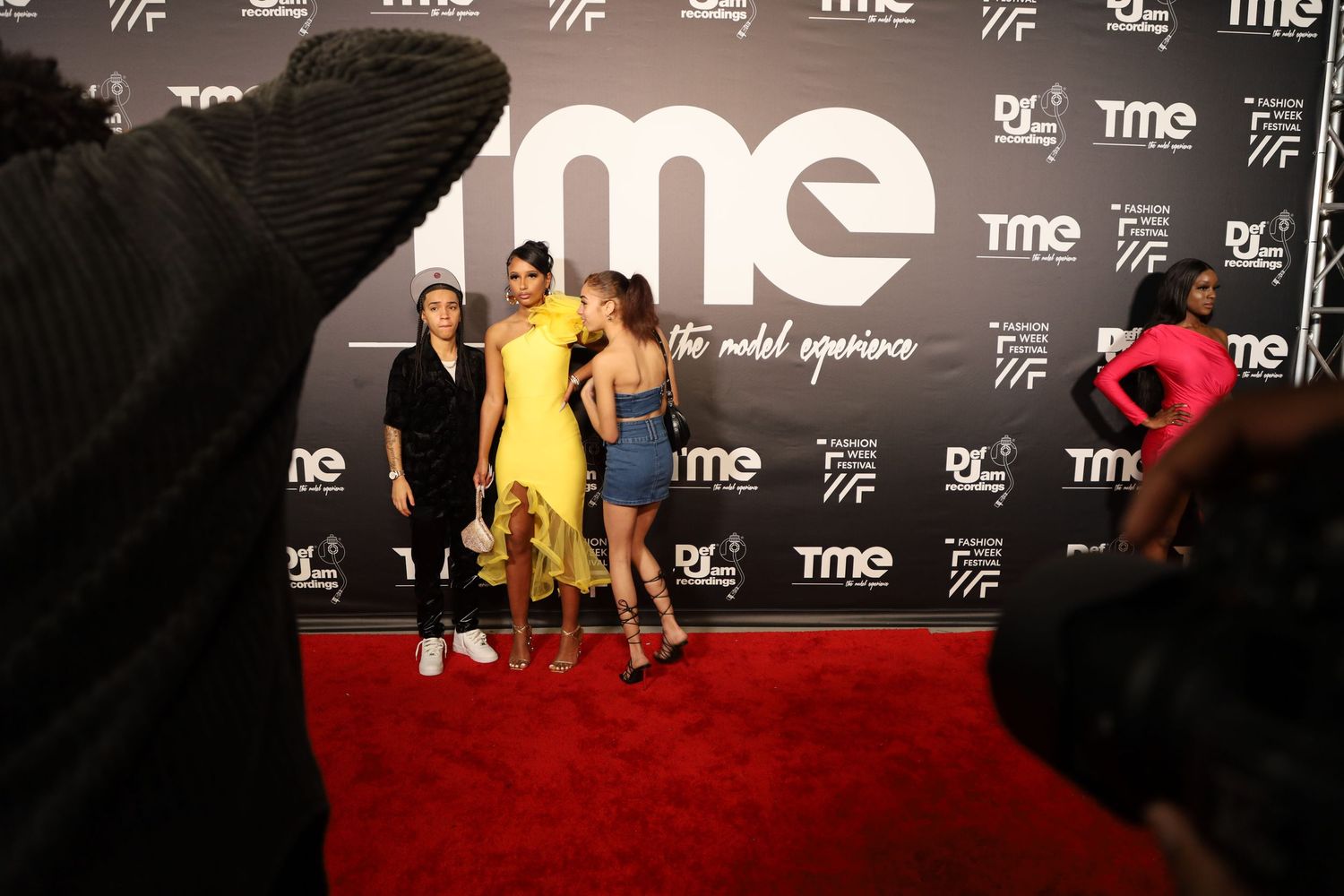 Fashion show attendees pose on red carpet in front of black TME branded backdrop.