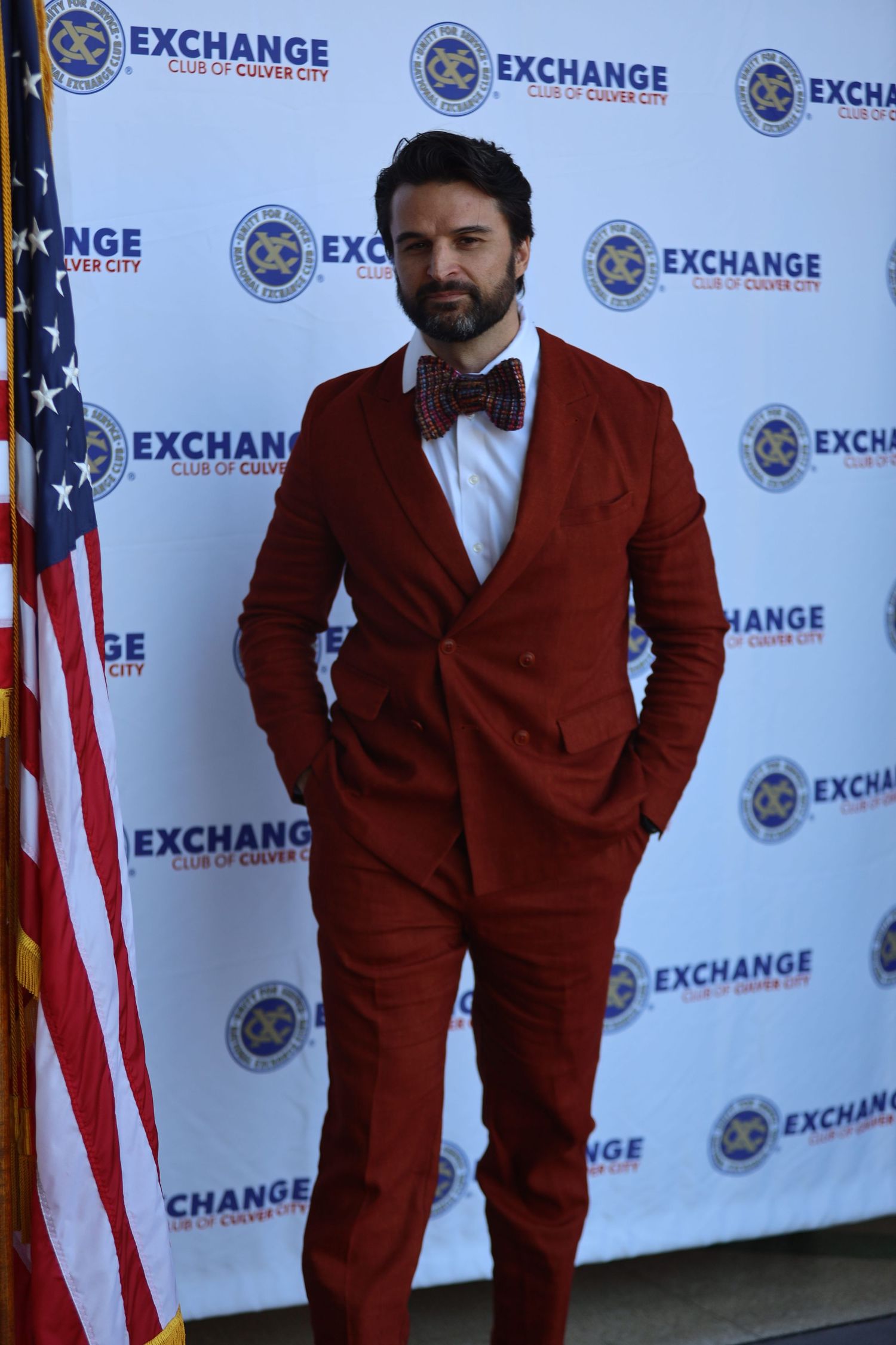 Person in dark red suit stands against Exchange logo backdrop at formal event.