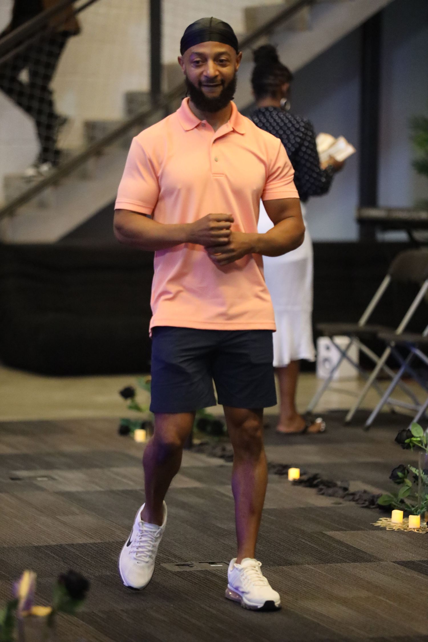 Person in coral polo shirt and navy shorts performing dance moves on wooden floor.