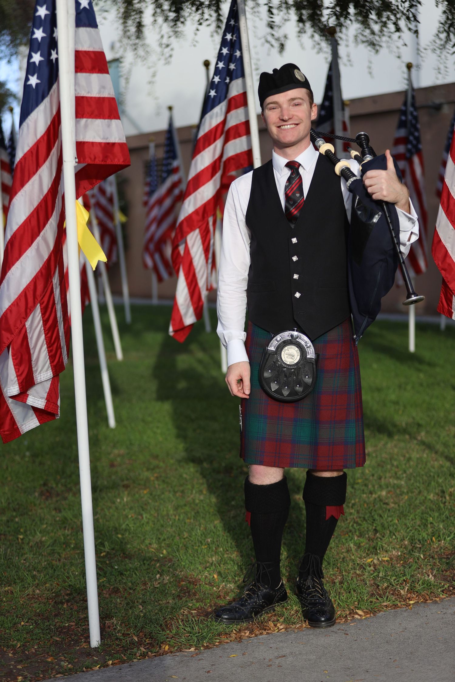 Scottish bagpiper in traditional dress stands among rows of American flags on grass.
