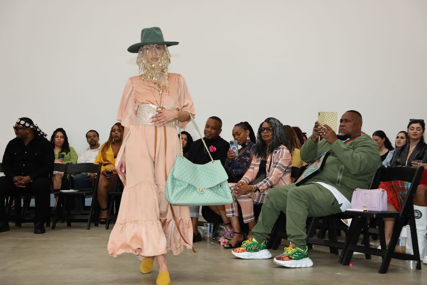 Model in pink dress and hat performs on runway with seated audience members visible.