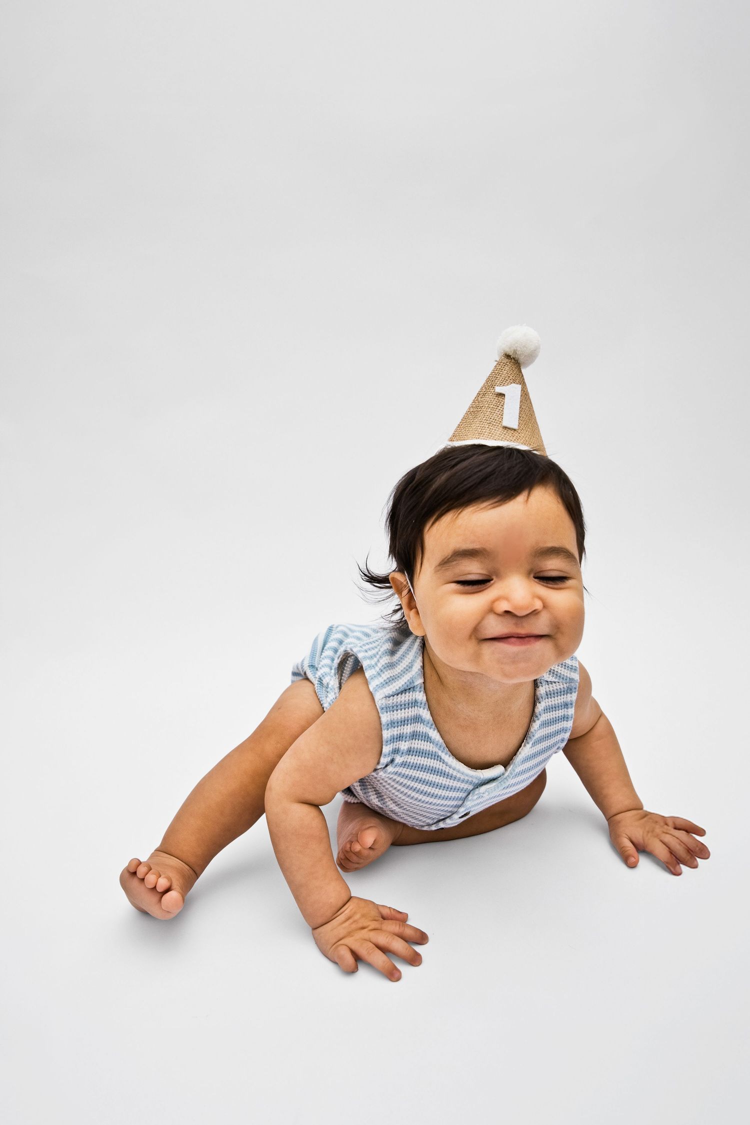 Smiling baby in striped onesie wearing a number 1 birthday party hat, crawling on white background.