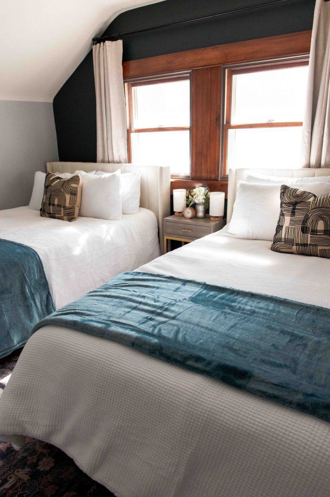 Two neatly made twin beds with teal velvet runners and decorative pillows in a cozy attic bedroom with dark walls.