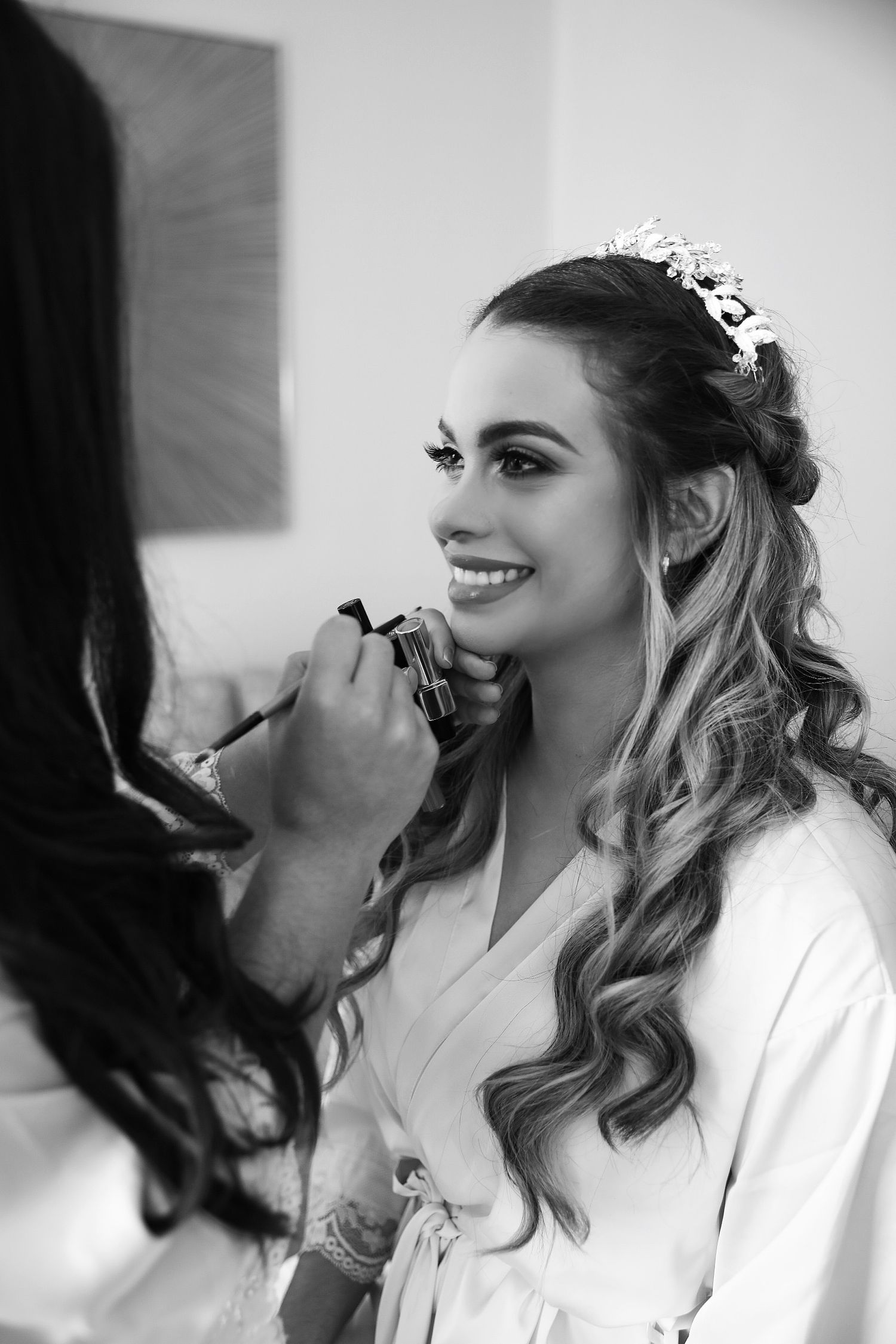 Black and white photo of wedding preparation with makeup being applied to a smiling bride wearing a white robe and tiara.
