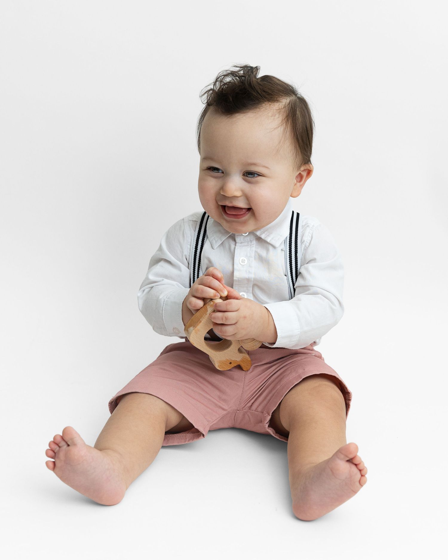 Baby in white shirt and pink shorts sits playfully on white background.