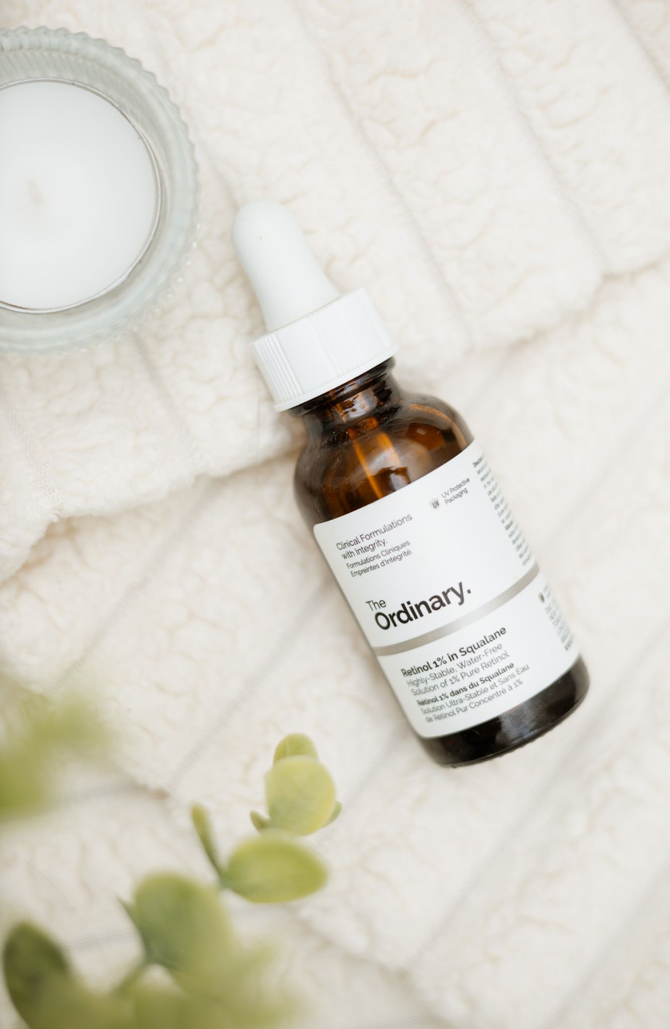 The Ordinary Retinol serum in amber dropper bottle on white knit fabric beside a candle and green plant.