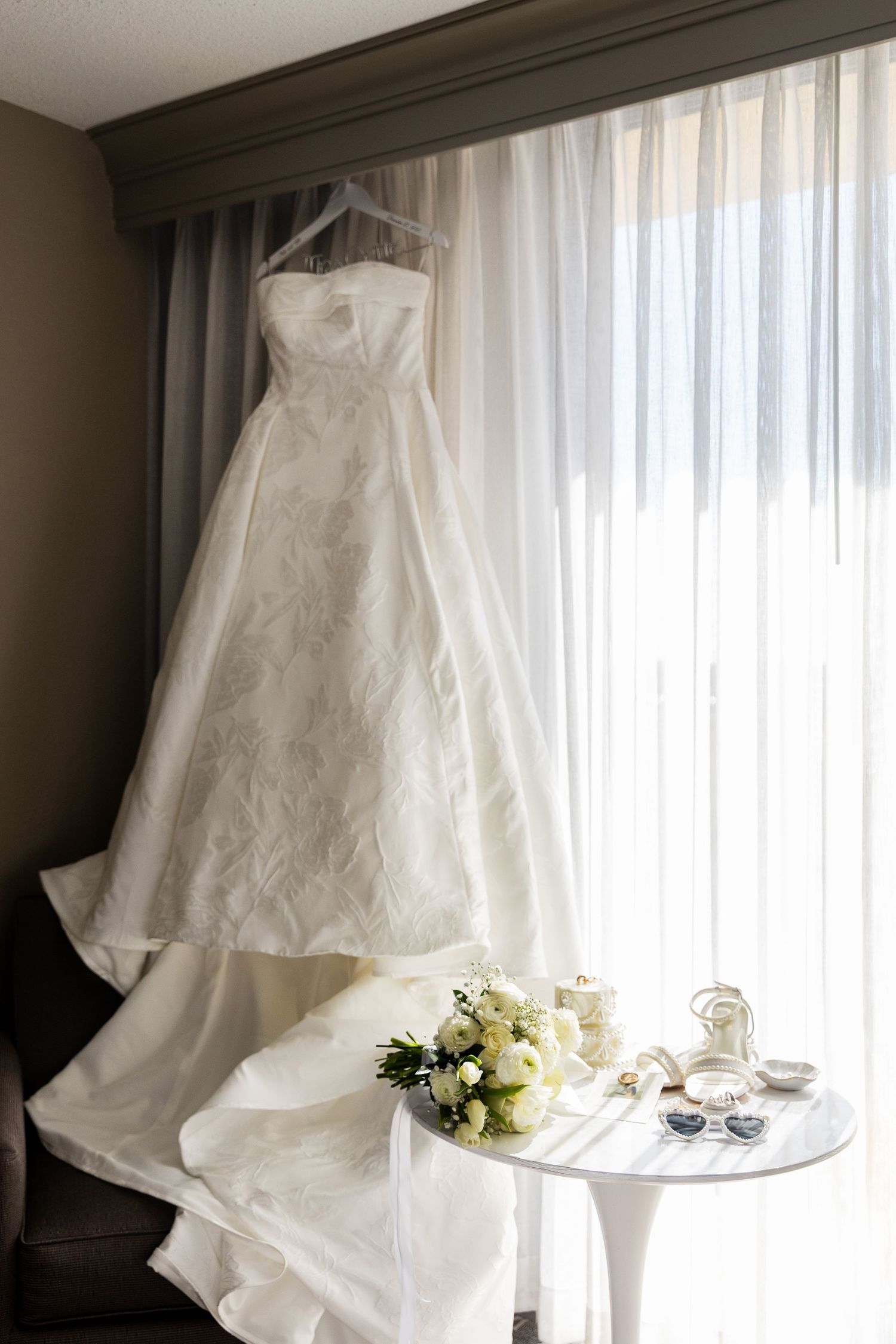 A white wedding dress hangs elegantly by a window with sunlight streaming through the delicate fabric.