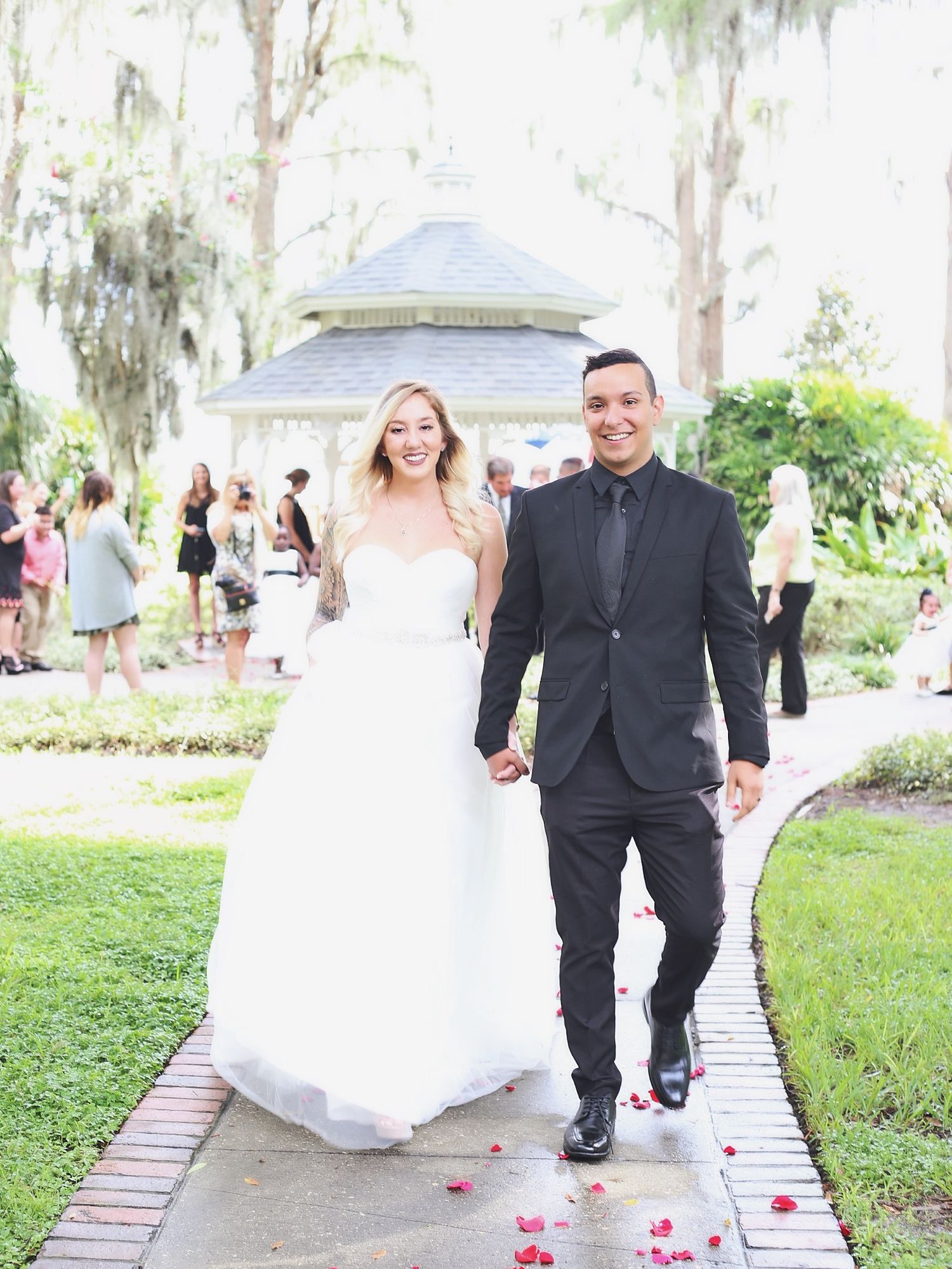 Newlyweds walking along a brick pathway with scattered rose petals at an outdoor garden venue.
