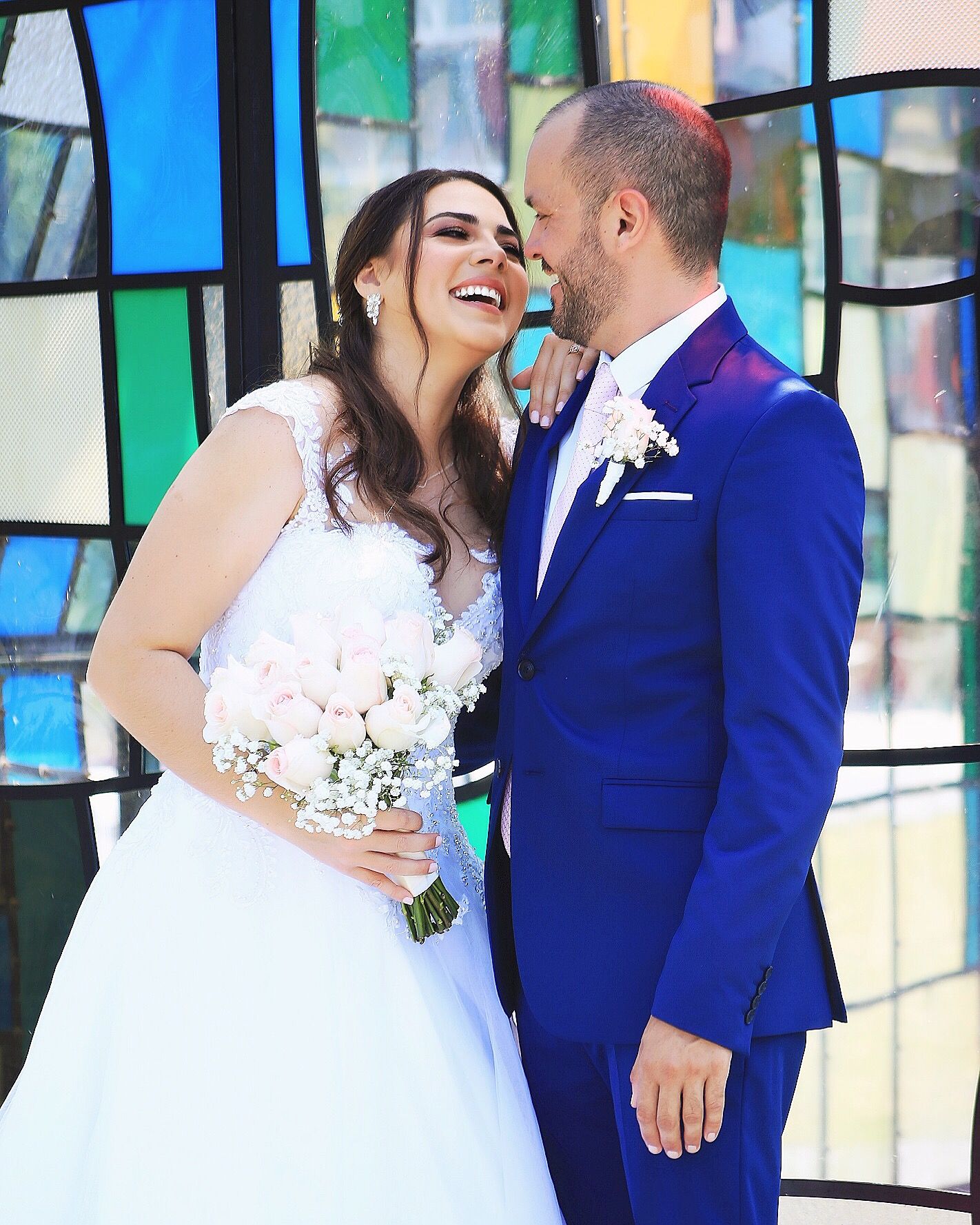 A couple shares a joyful moment in front of a colorful stained glass window backdrop on their wedding day.