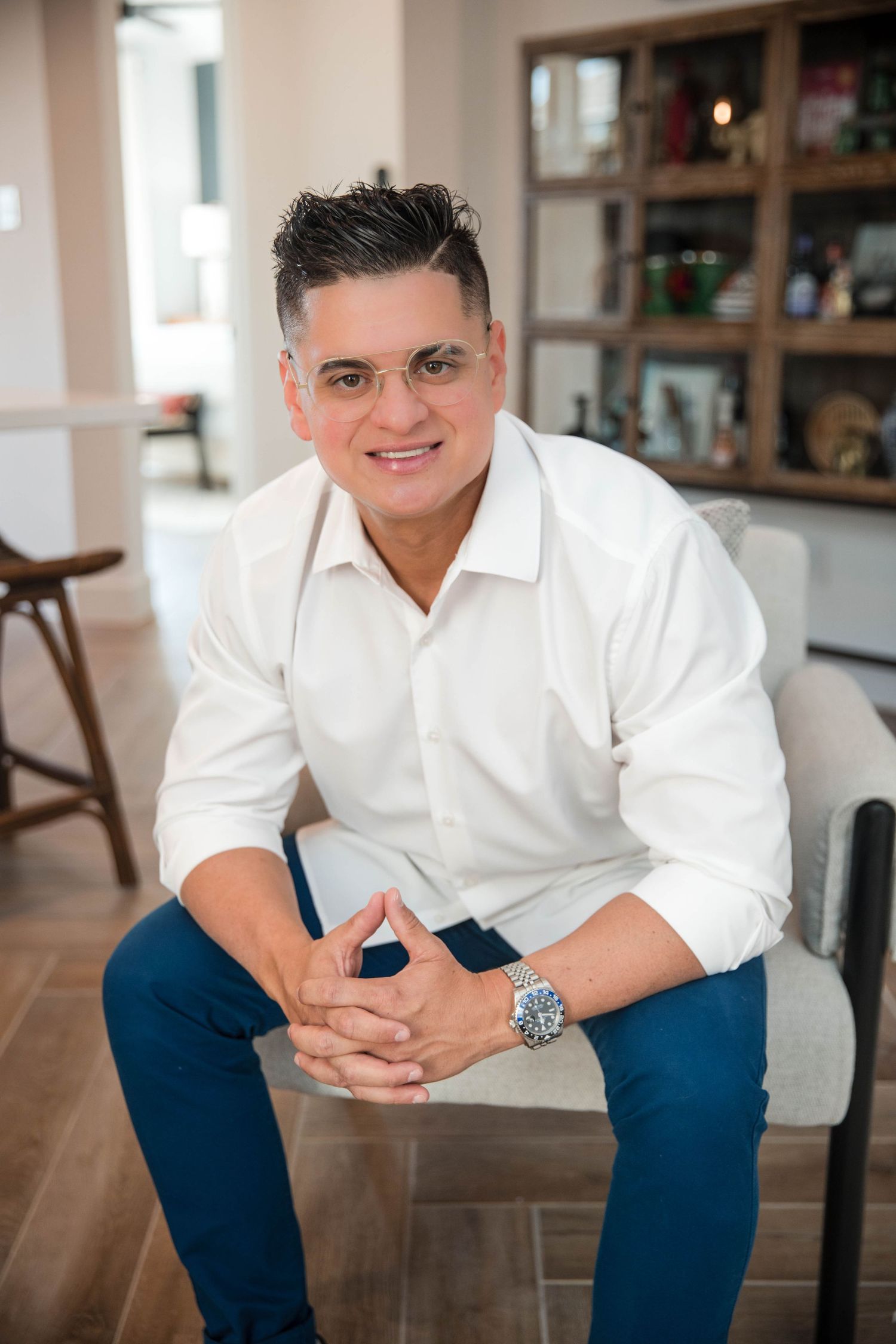 Professional portrait of person in white button-down shirt and navy pants sitting casually in modern indoor setting.